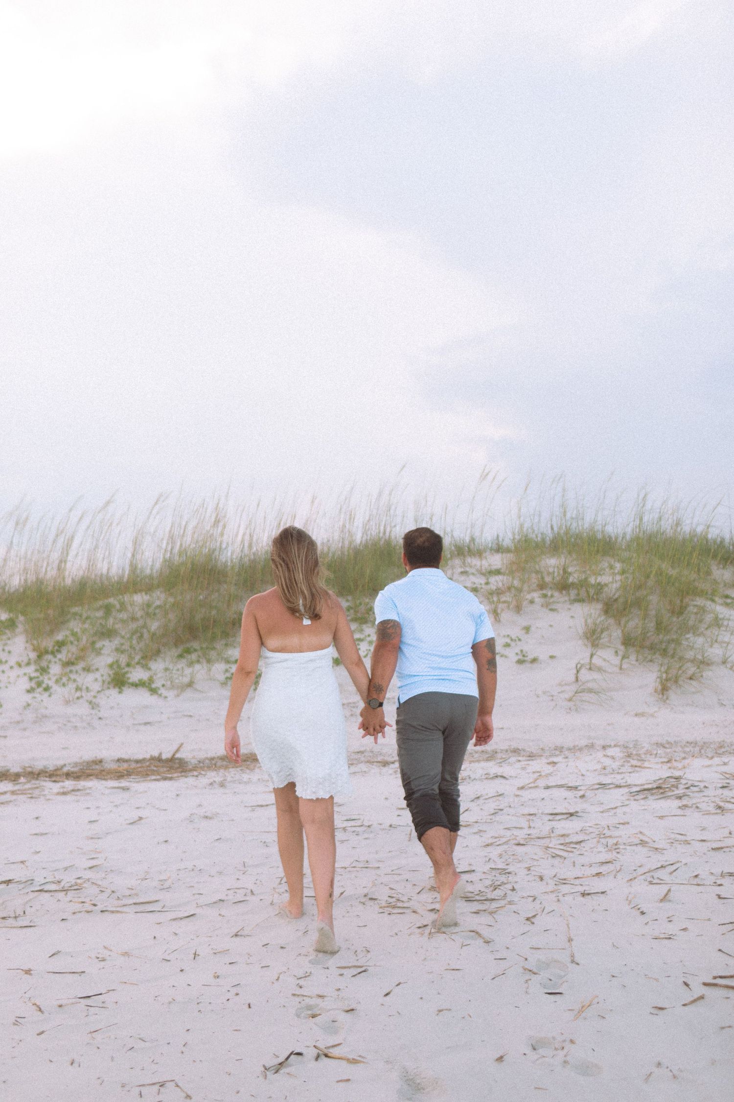 Couple walking hand in hand barefoot on a white sandy beach toward grass dunes at dusk.