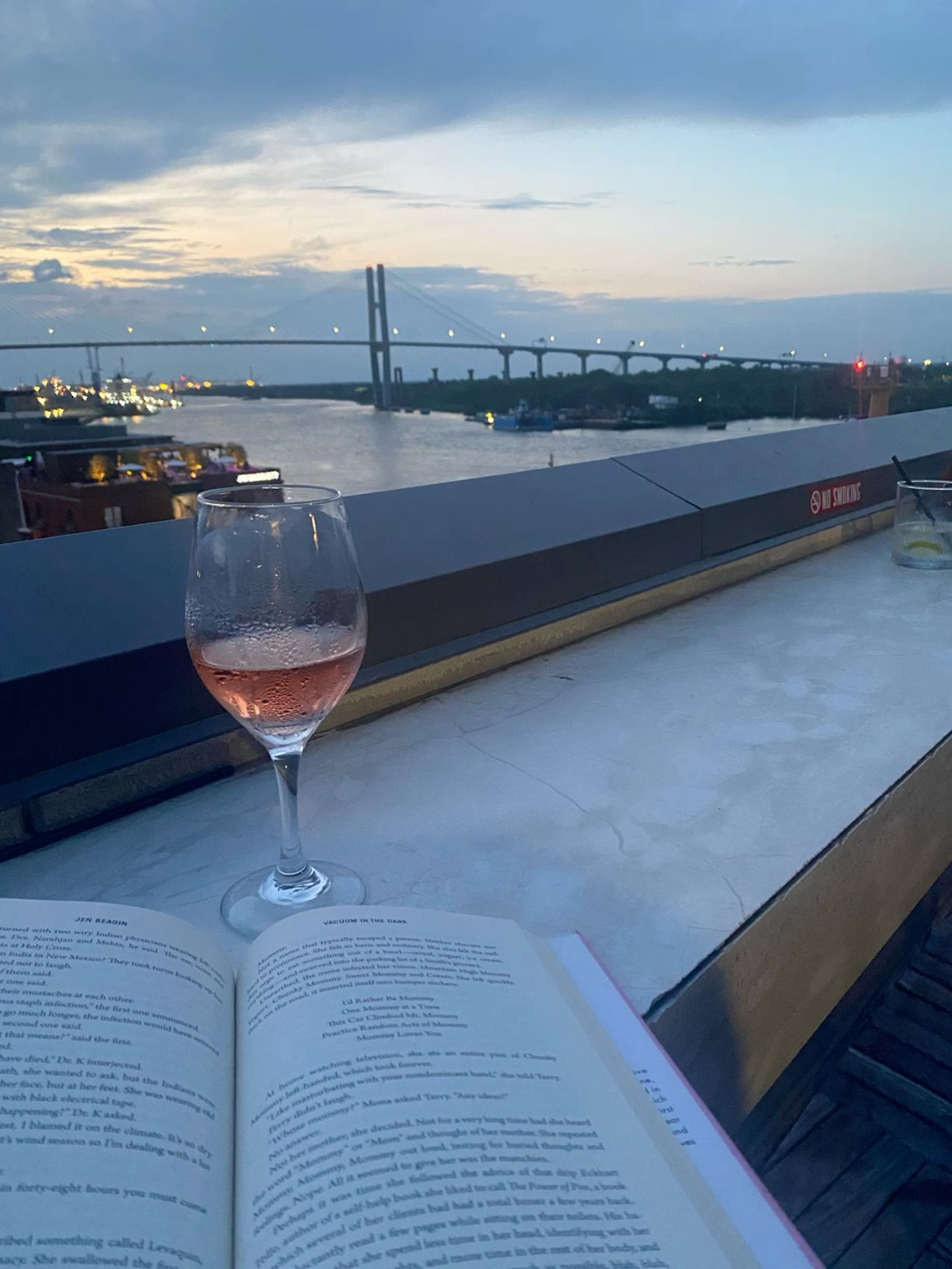 Glass of rose wine beside open book on rooftop bar at sunset with bridge views.