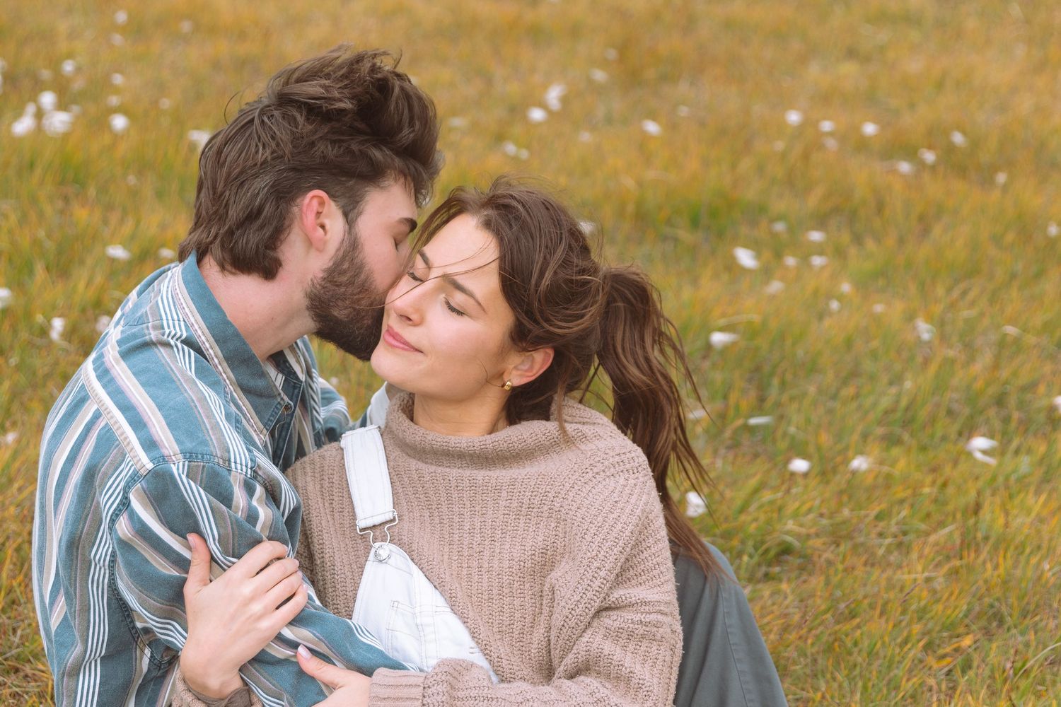 Bearded man kissing woman on cheek while sitting together in a flower-dotted meadow.
