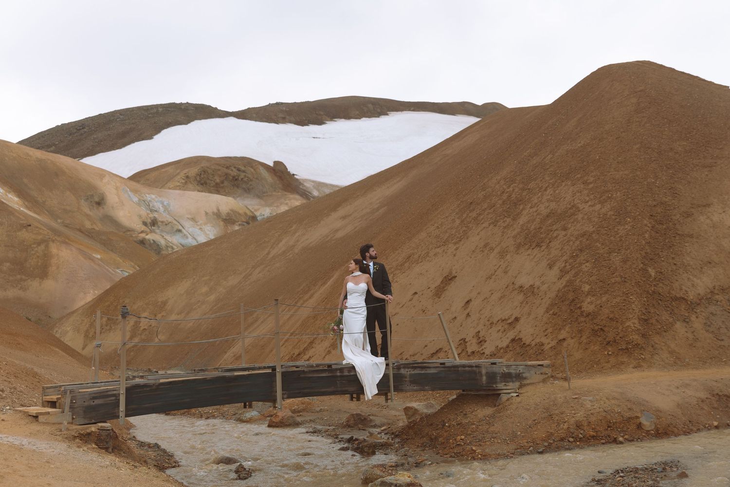 Wedding couple stands close together on a bridge surrounded by golden volcanic hills in Iceland.