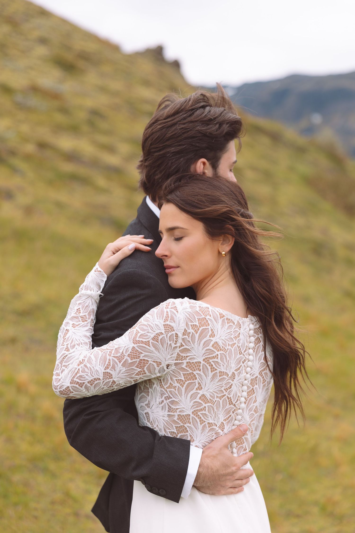 Bride with flowing hair embraces groom tightly against green hillside in outdoor wedding shoot.