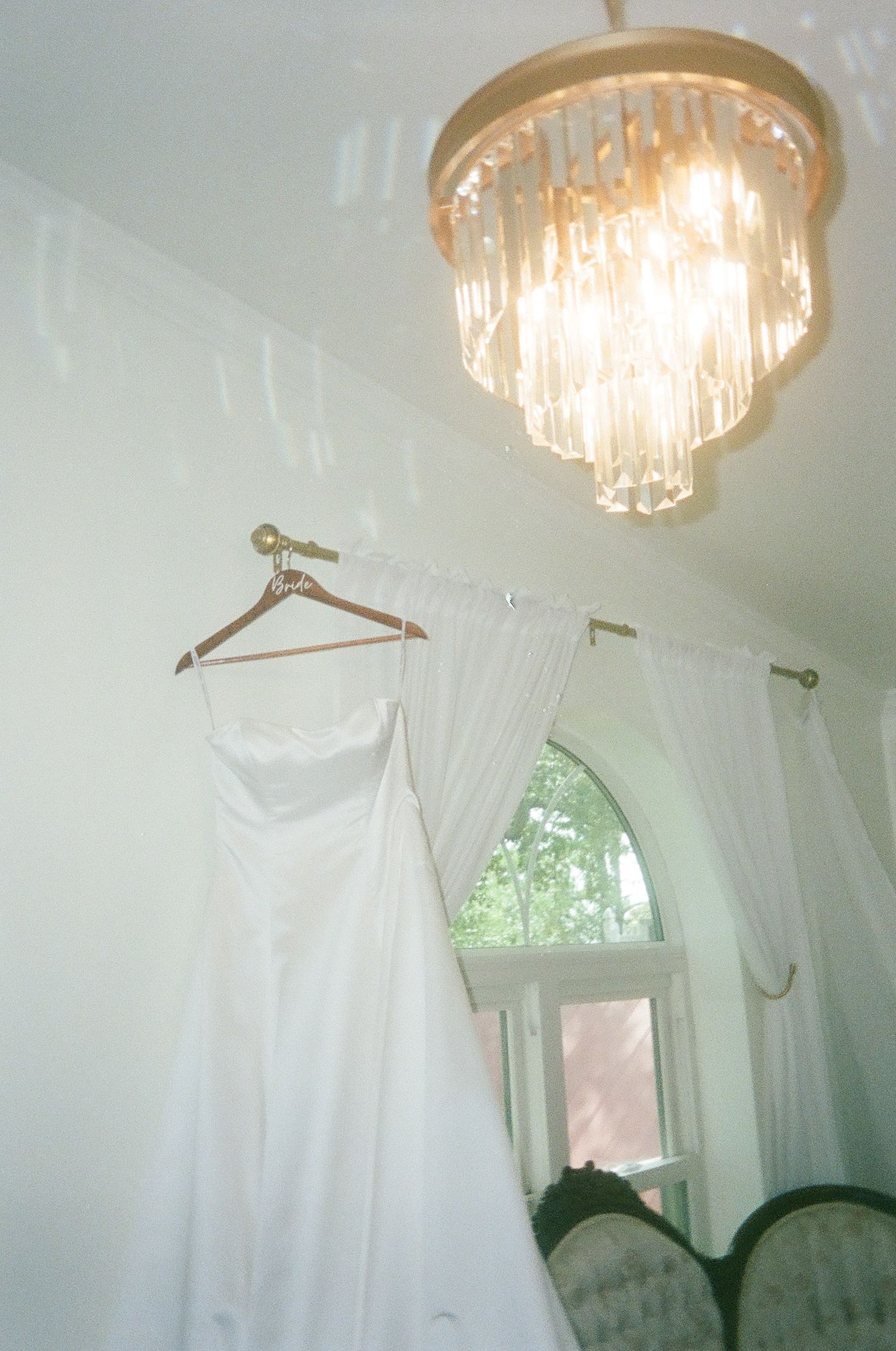 White wedding dress on wooden hanger near arched window with crystal chandelier in elegant bridal suite.