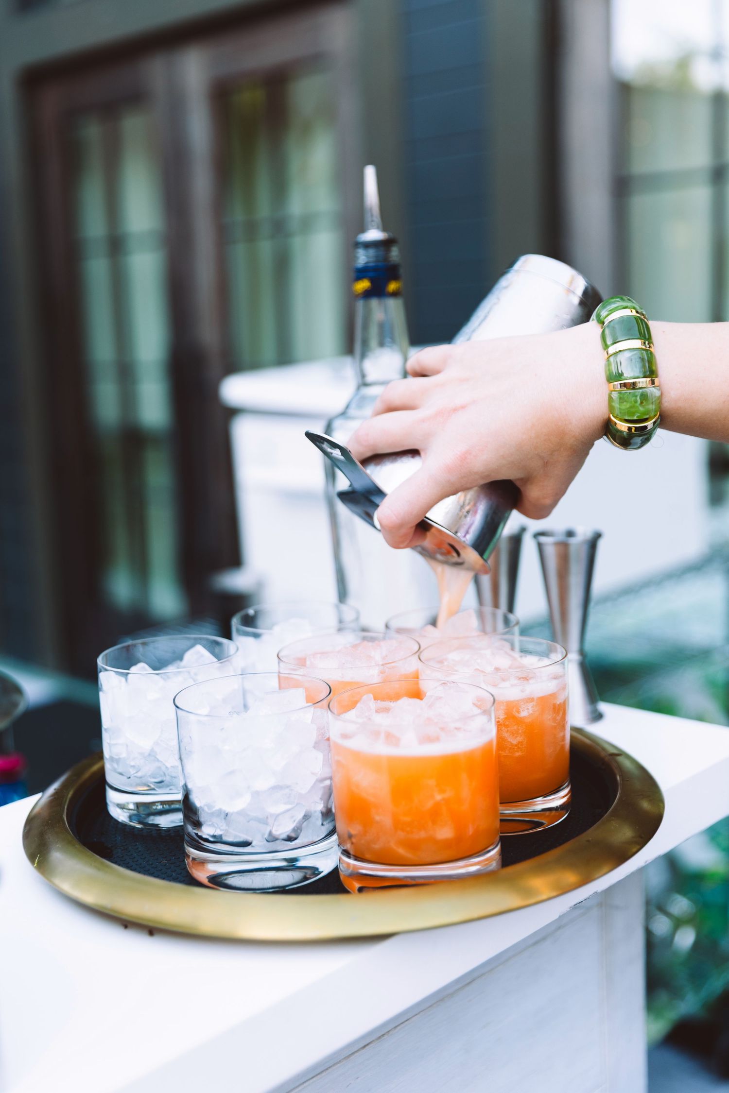 Bartender pouring orange cocktails into rocks glasses filled with ice on a gold tray at an outdoor bar.