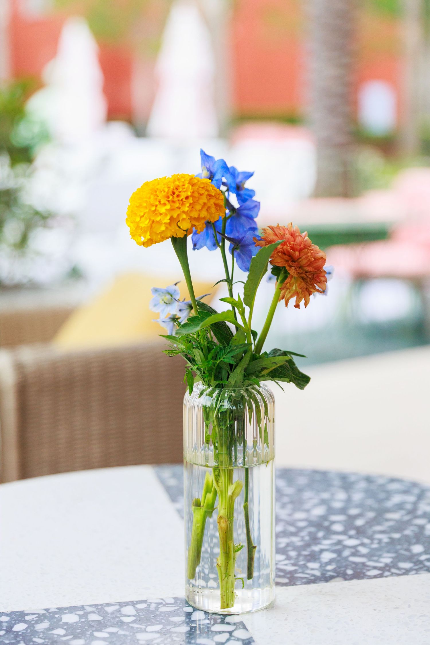 Colorful bouquet of marigolds, blue flowers, and orange dahlias in a clear glass vase on an outdoor patio table.