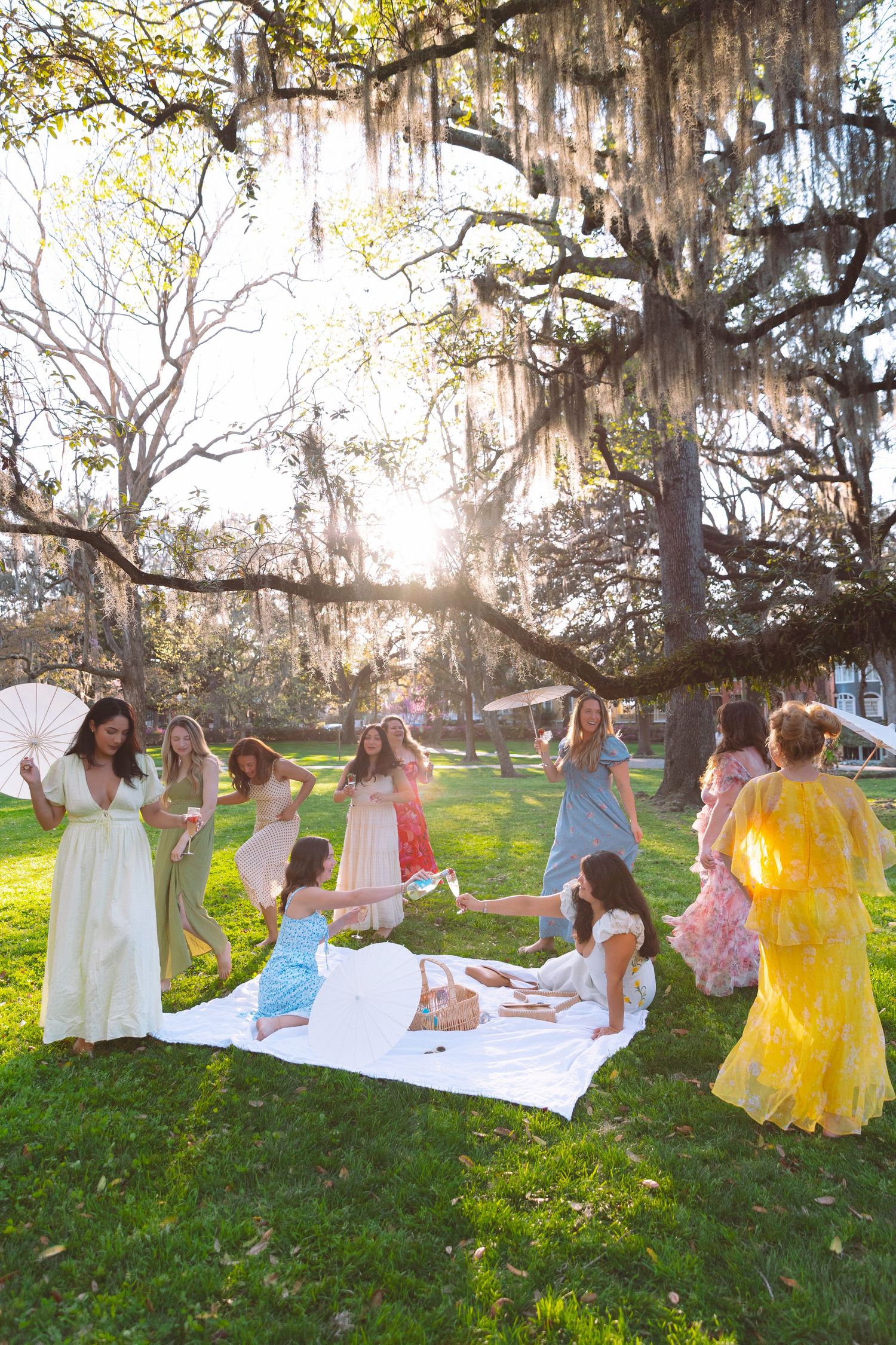Women in pastel dresses gather outdoors under Spanish moss-draped oak trees for a sunny garden party picnic.