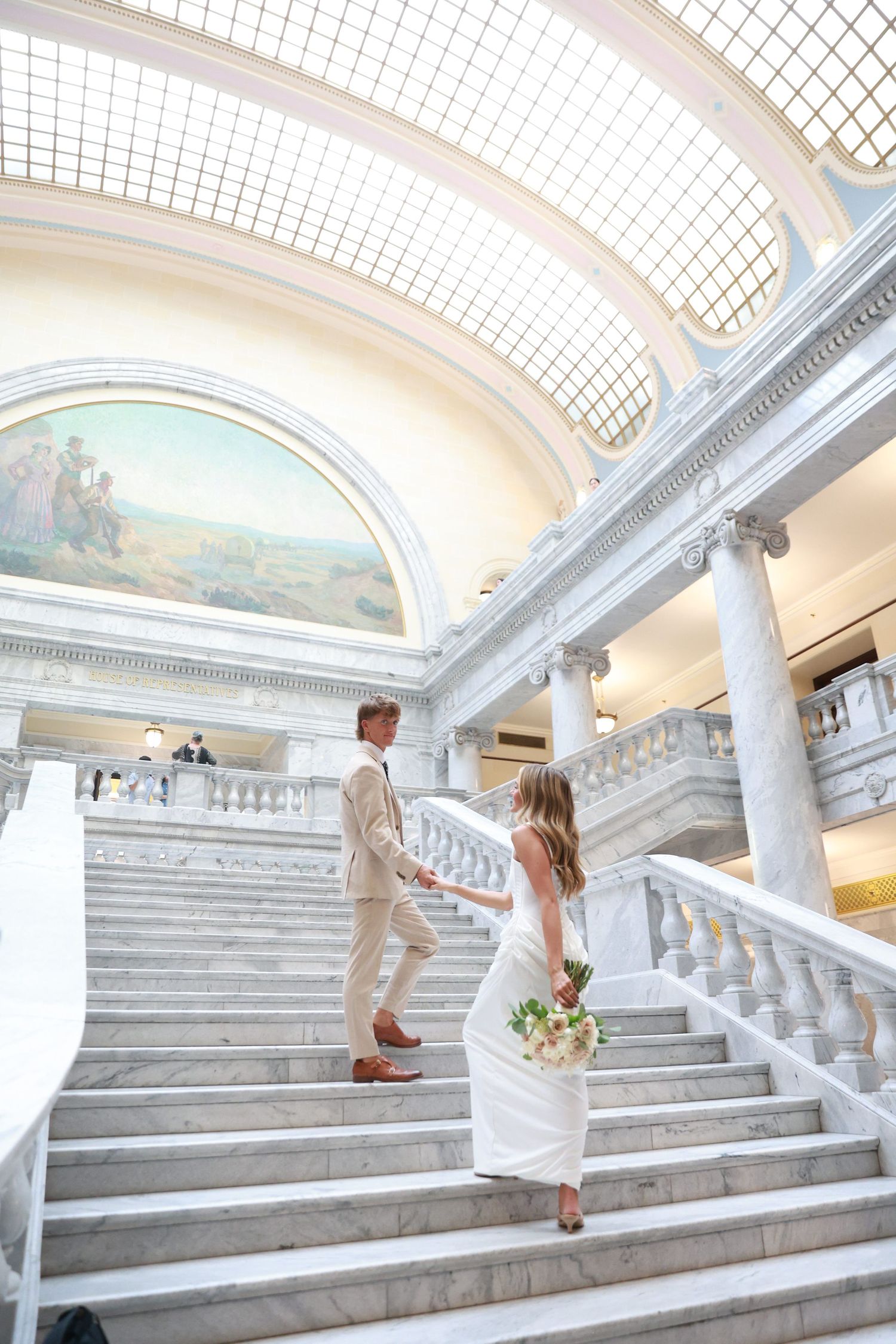 Bride and groom walking up grand marble staircase in elegant domed building, bride holding floral bouquet.