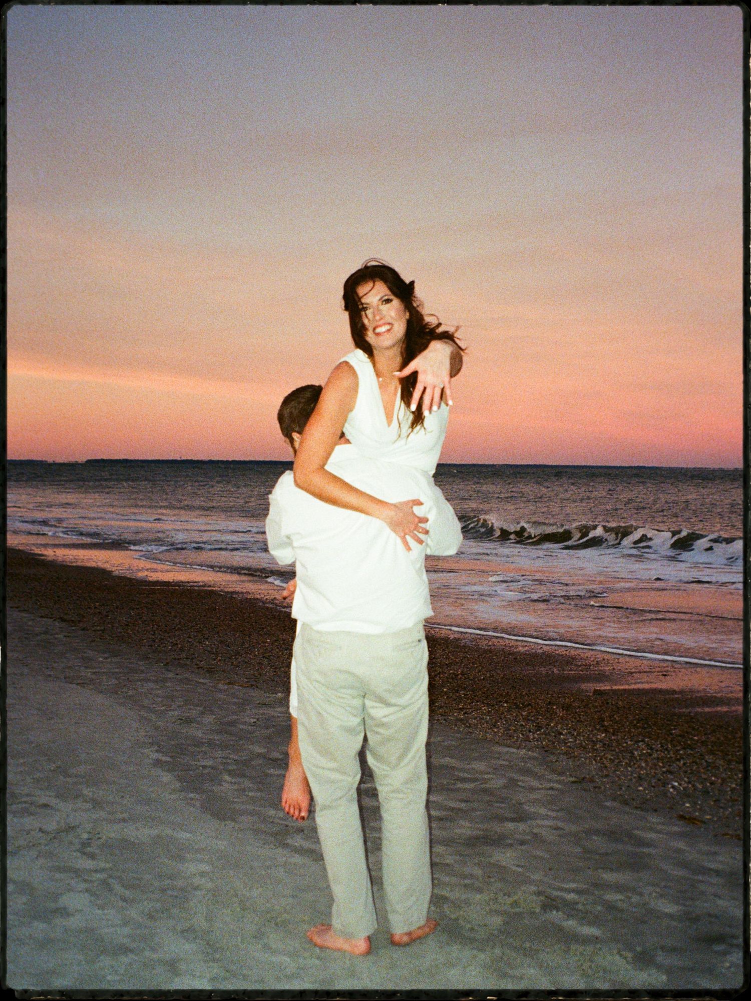 Man lifting woman in white dress on a beach during a colorful pink and purple sunset.