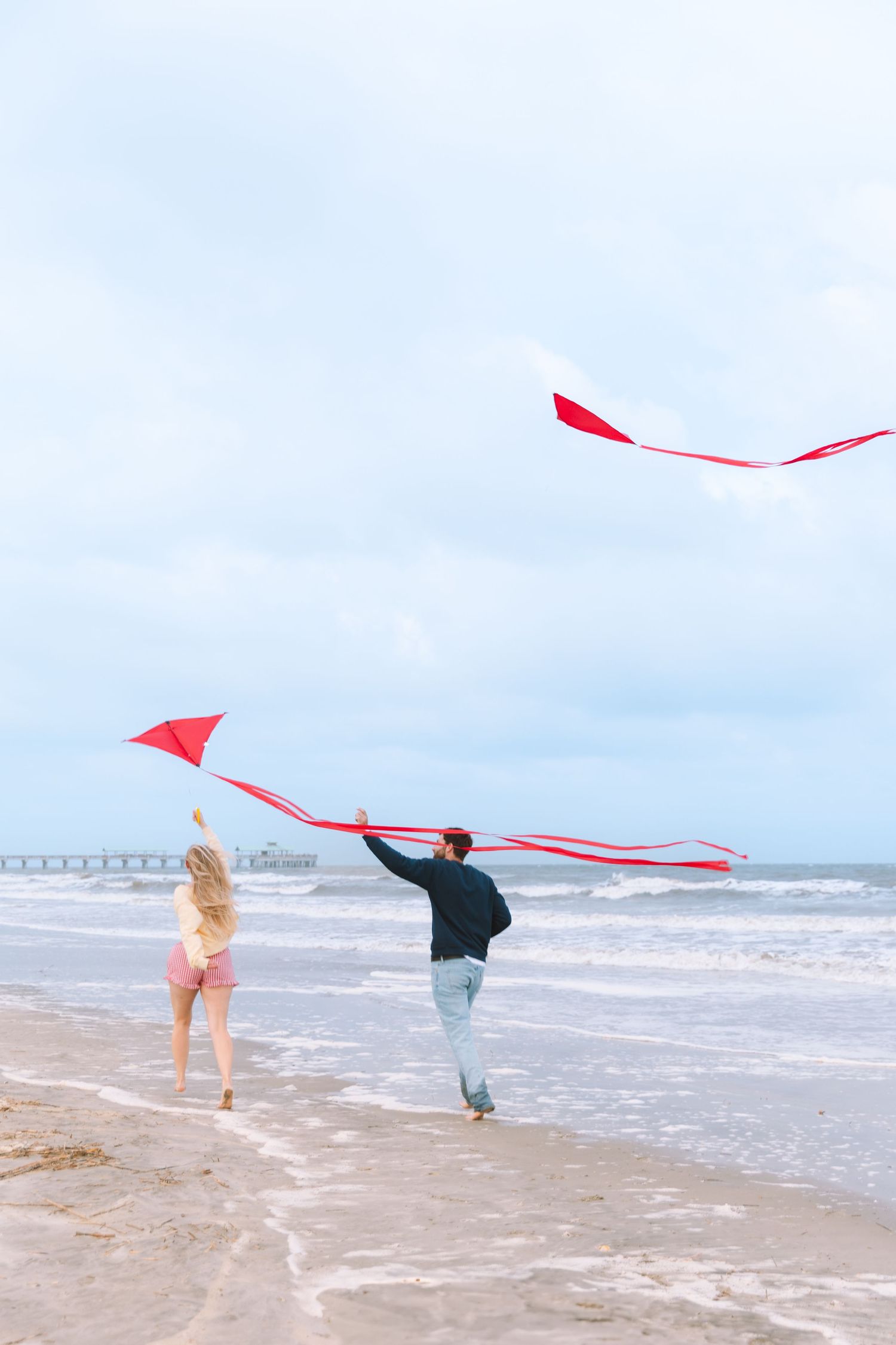 Couple flying red kites together on a cloudy beach near ocean waves and a distant pier.