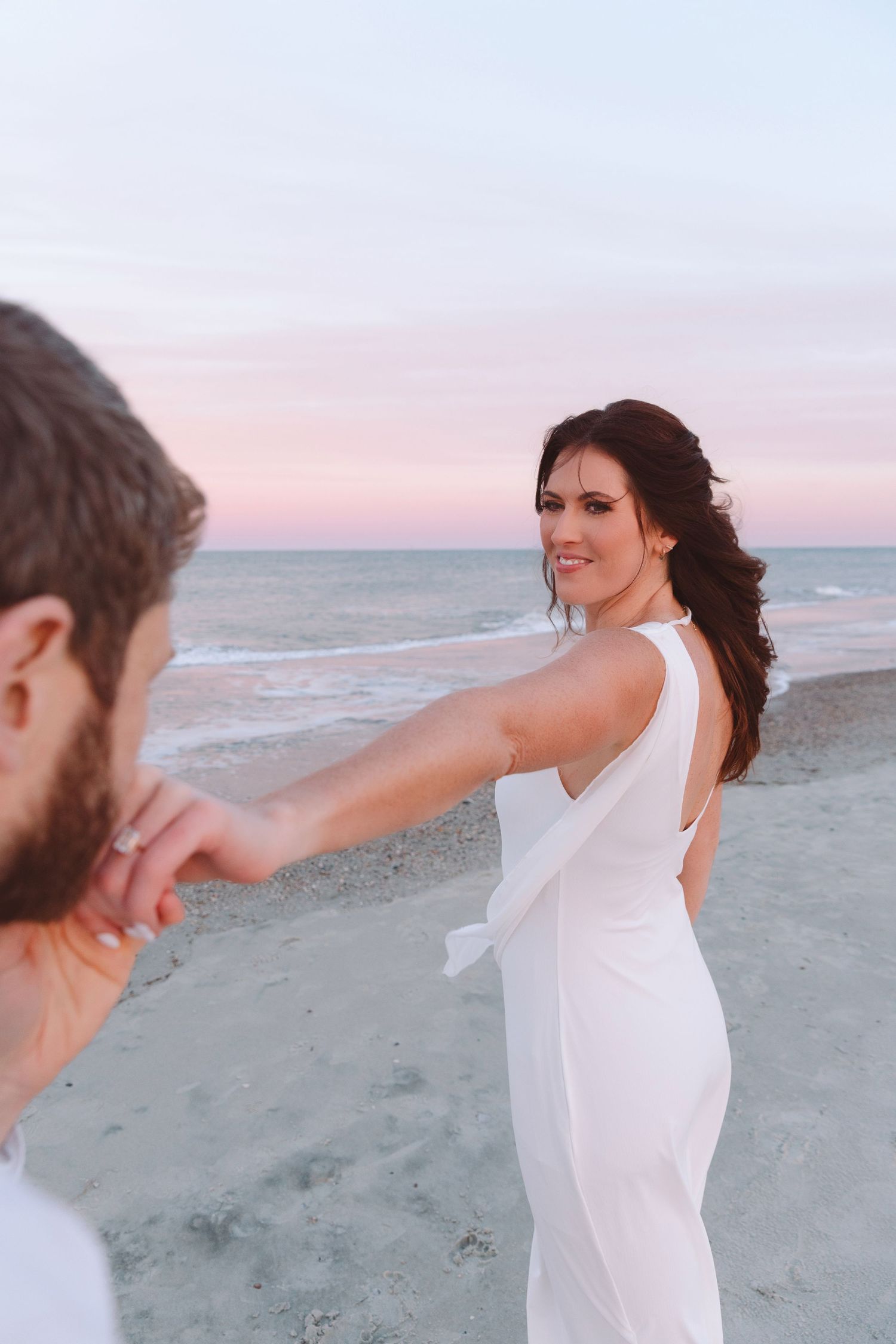 A bride in a white dress holds her groom's hand on a beach at sunset.