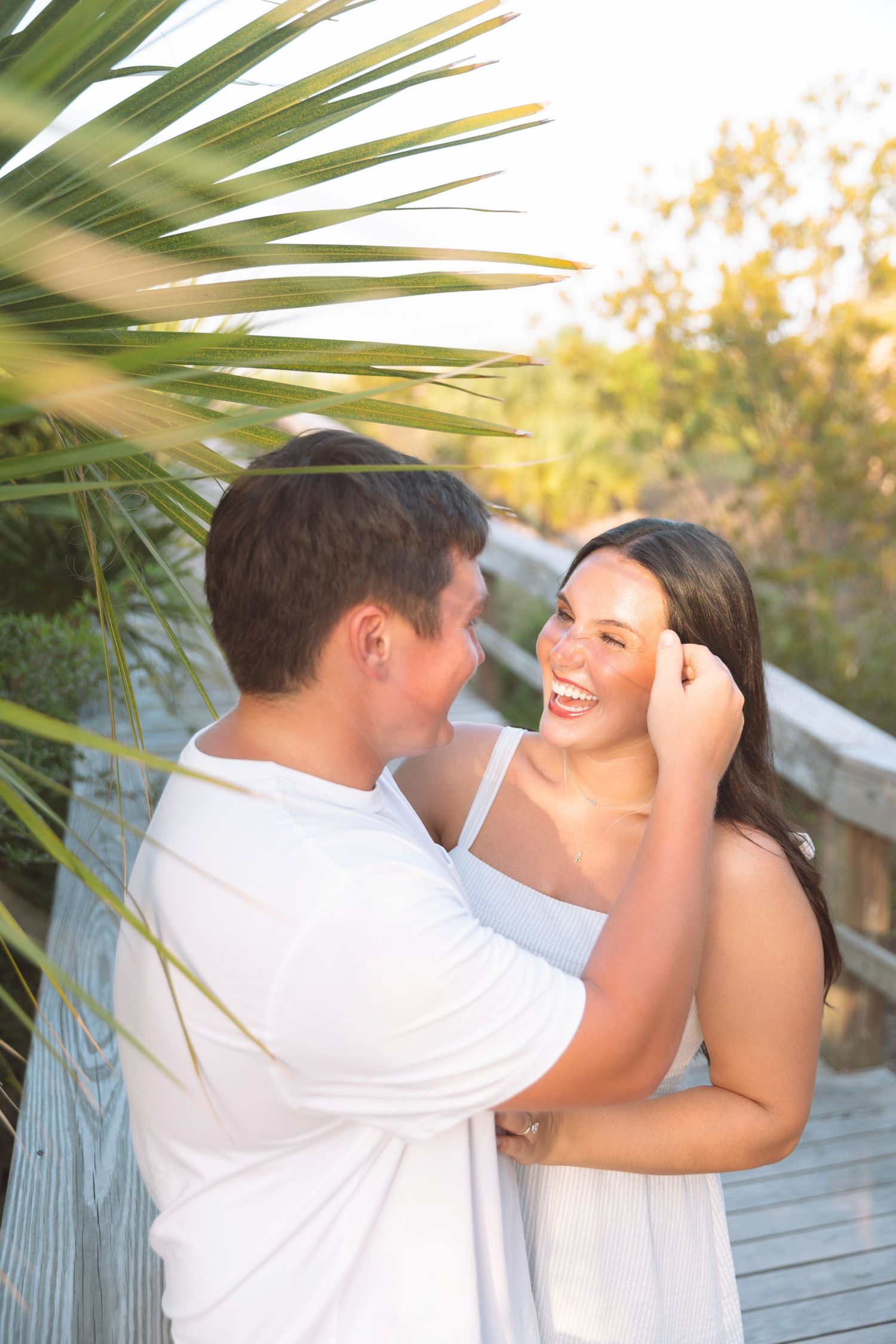 Happy couple in white outfits sharing a romantic moment near a palm tree on a sunny wooden boardwalk.