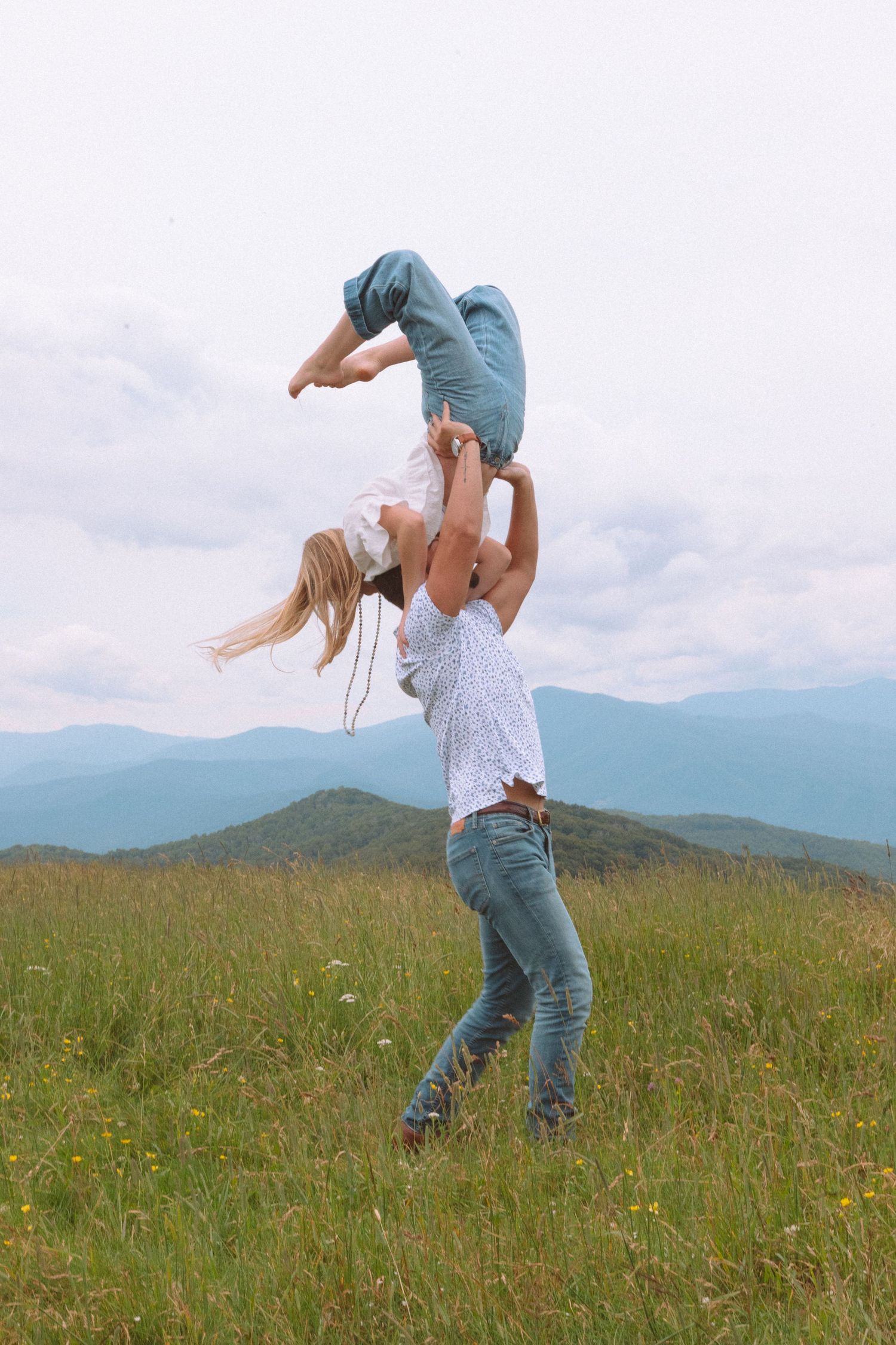 Man lifting woman overhead in a grassy mountain meadow under cloudy skies.