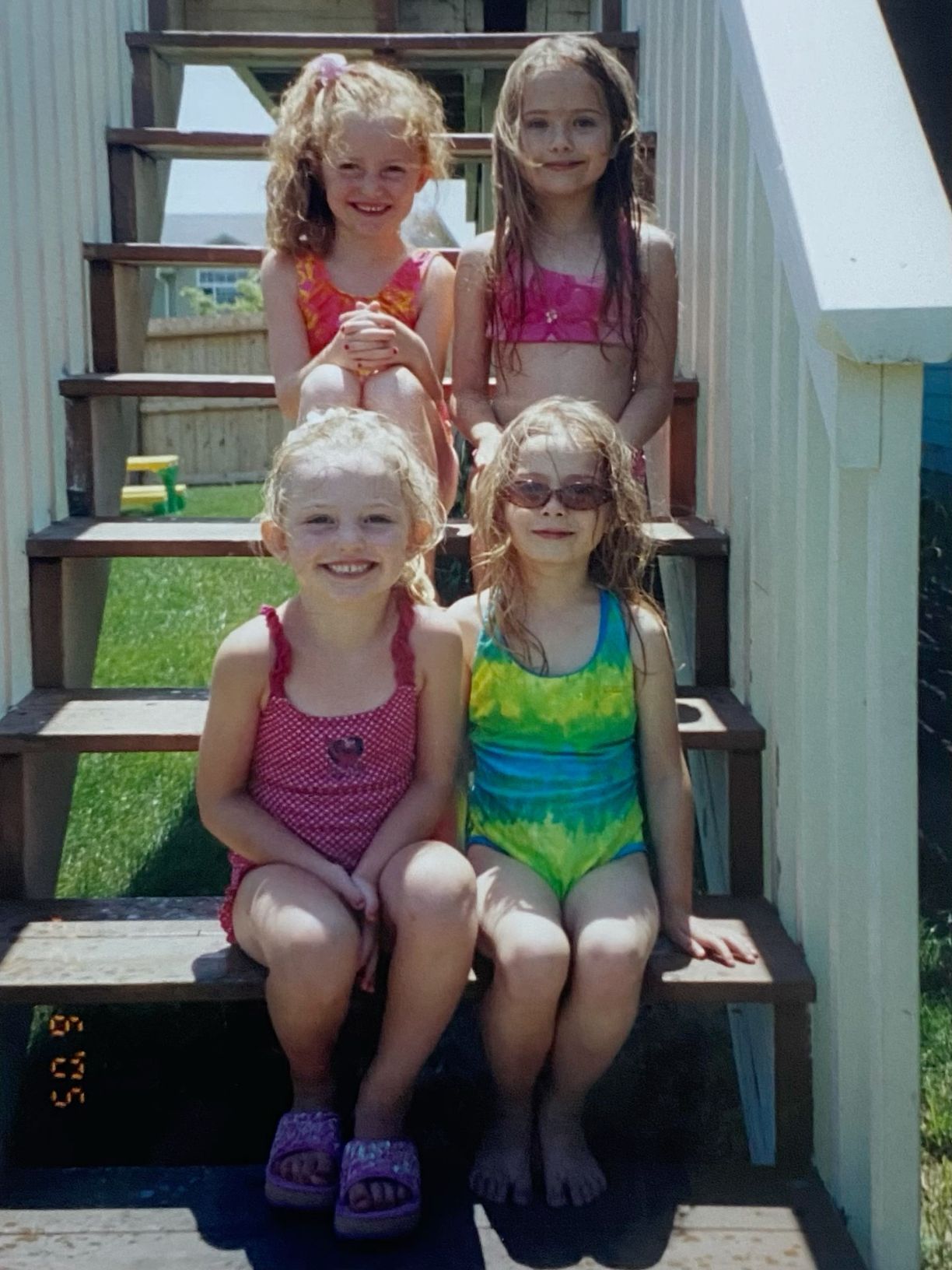 Four young girls in colorful swimsuits posing on wooden outdoor steps on a sunny summer day.
