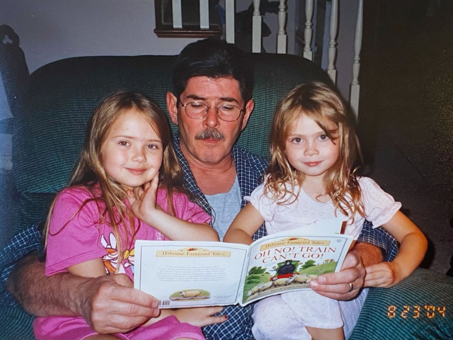Grandfather reading a colorful picture book with two young granddaughters on a cozy armchair.