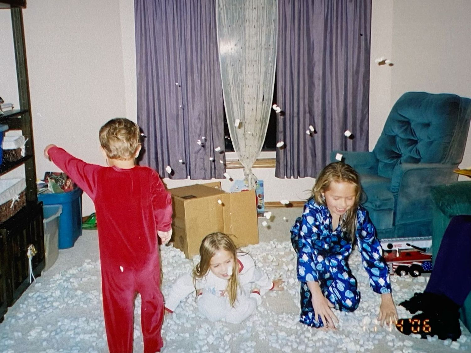 Three children in pajamas play in a living room covered with scattered white packing peanuts.