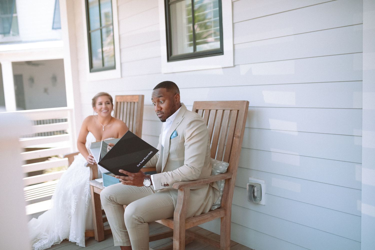 Groom in tan suit reading a book on porch bench while smiling bride sits nearby.