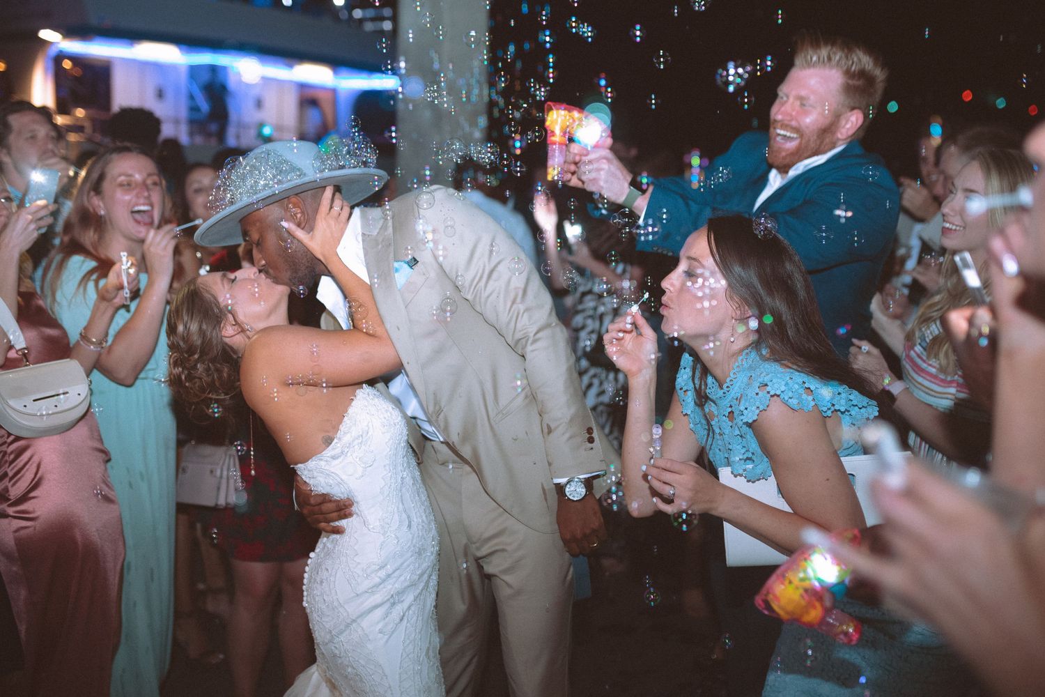 Bride and groom kiss during festive bubble send-off at outdoor nighttime wedding.