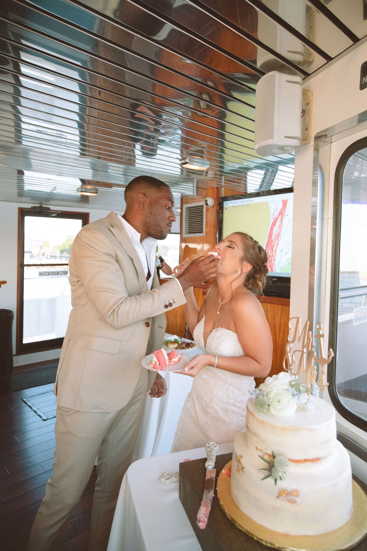 Newlyweds share cake feeding moment beside two-tier wedding cake during boat reception celebration.