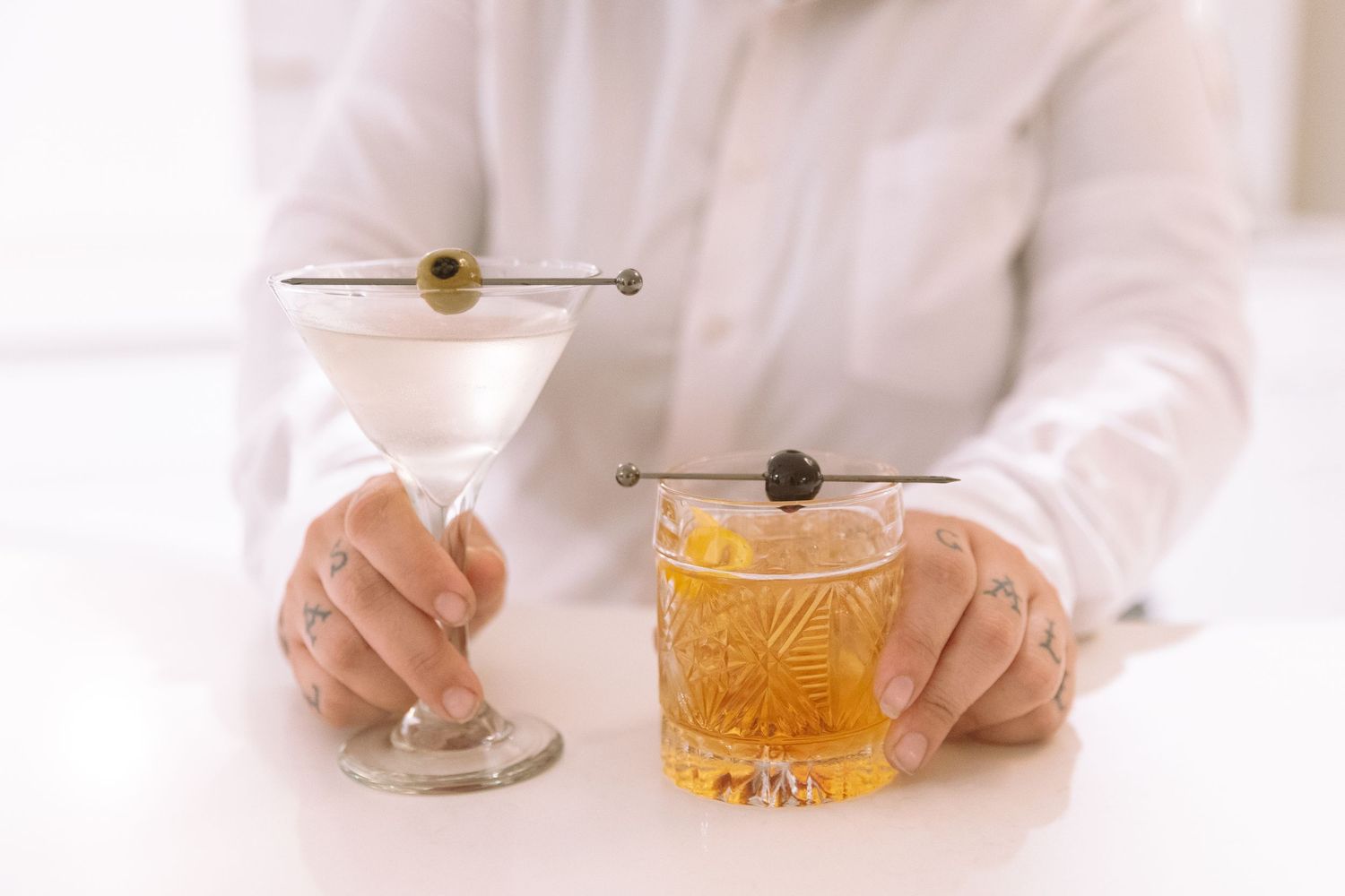 Two elegant cocktails, a dirty martini and an old fashioned, displayed on white bar surface.