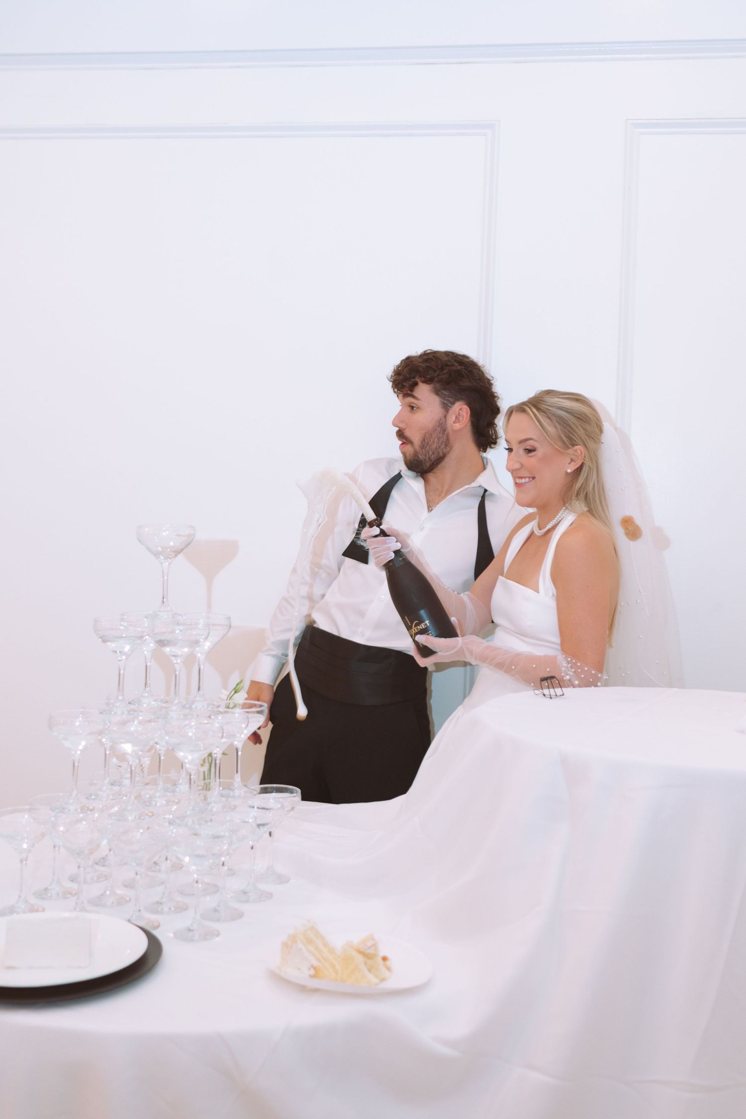 Bride and groom pouring champagne together at a coupe glass tower at their white wedding reception.