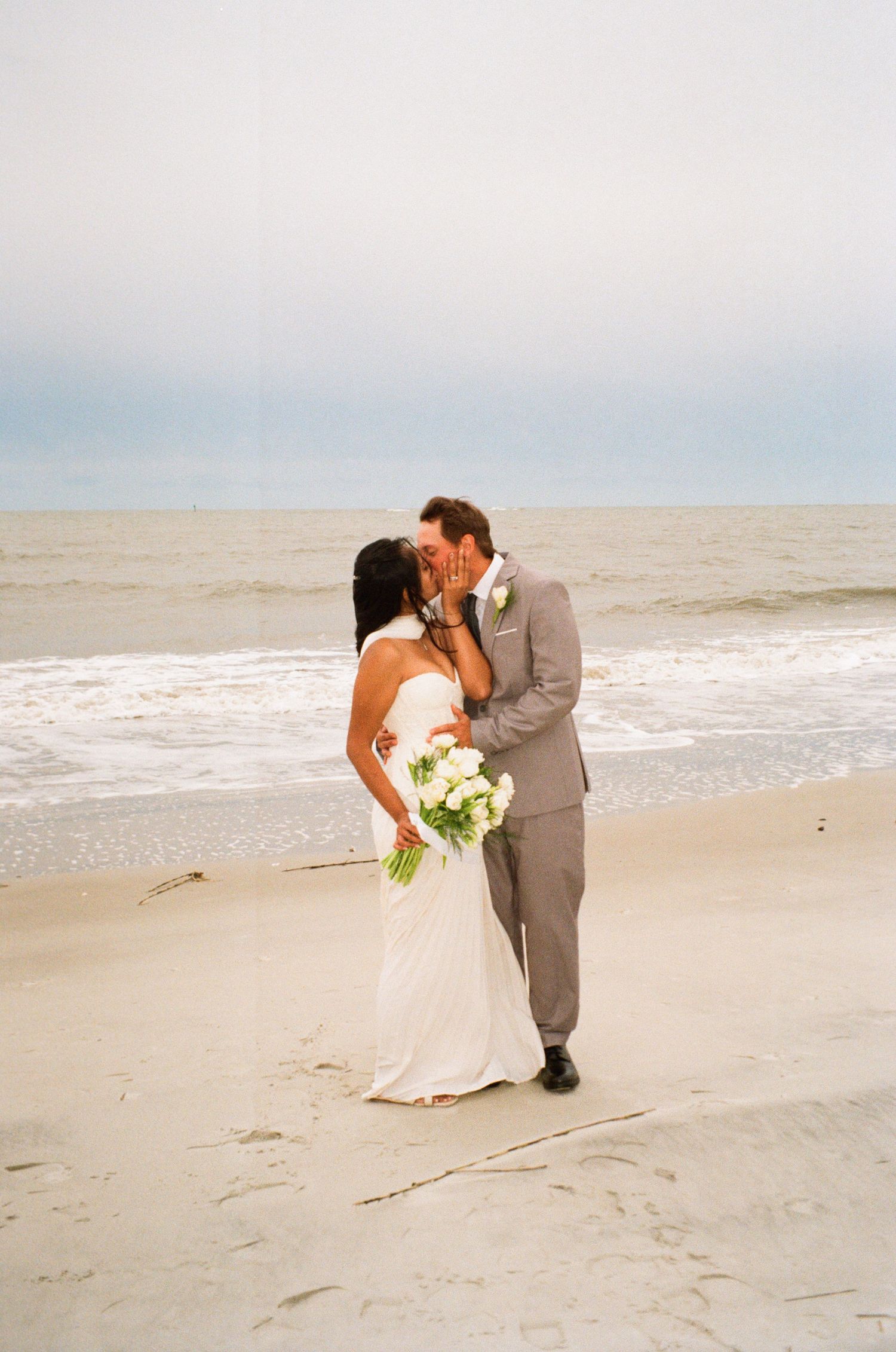 Bride and groom share a romantic beach kiss, she holds white bouquet, he wears gray suit, ocean waves in background.