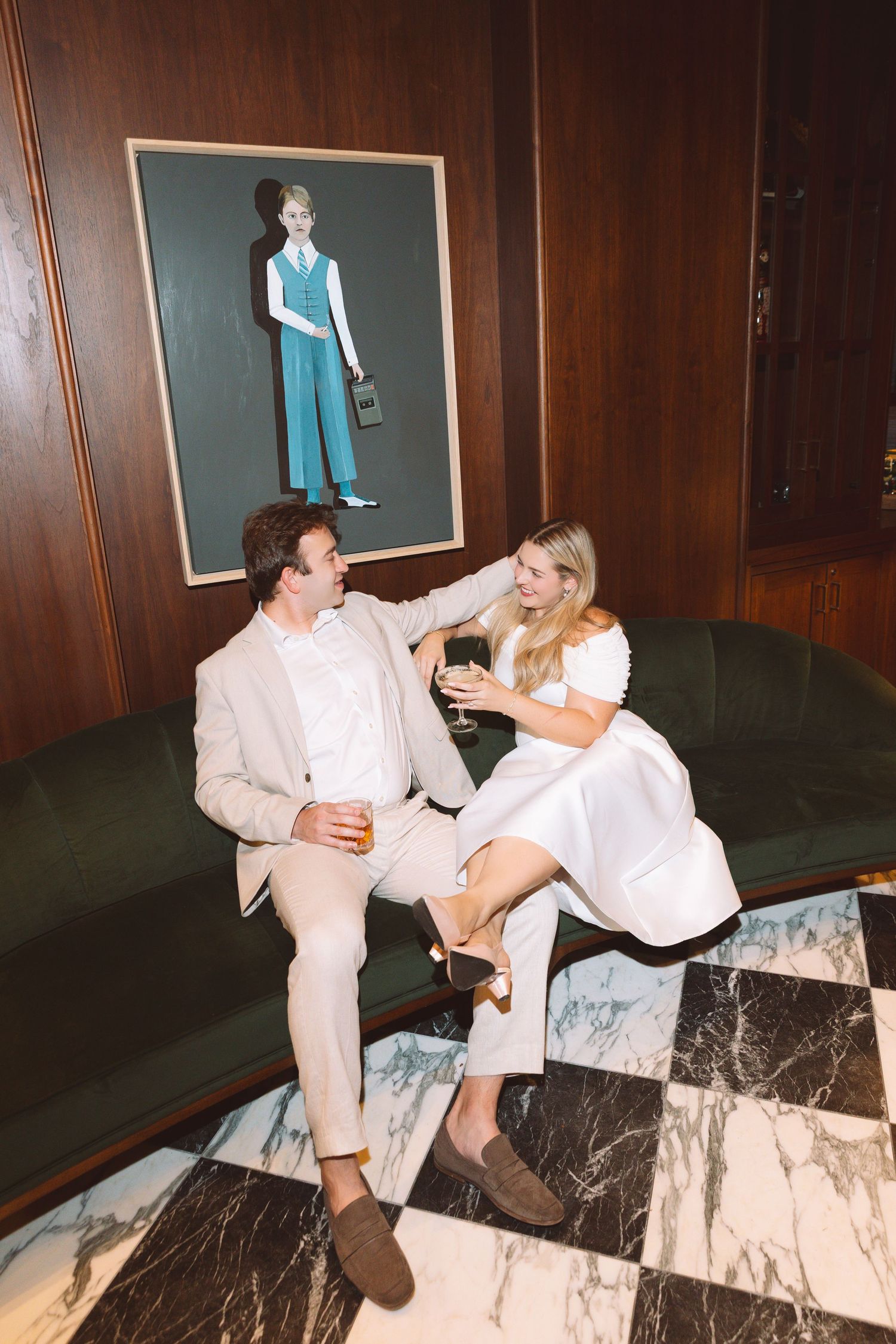 Couple in elegant attire toasting drinks on a green velvet sofa beneath a portrait painting in a sophisticated lounge.