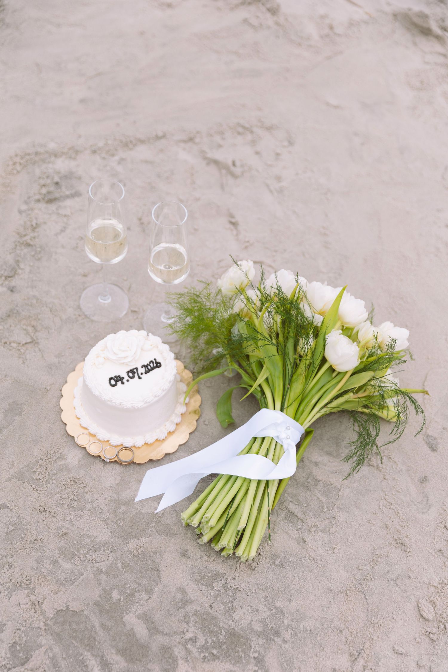 Beach wedding flat lay with small white cake, champagne glasses, and white tulip bouquet on sand.