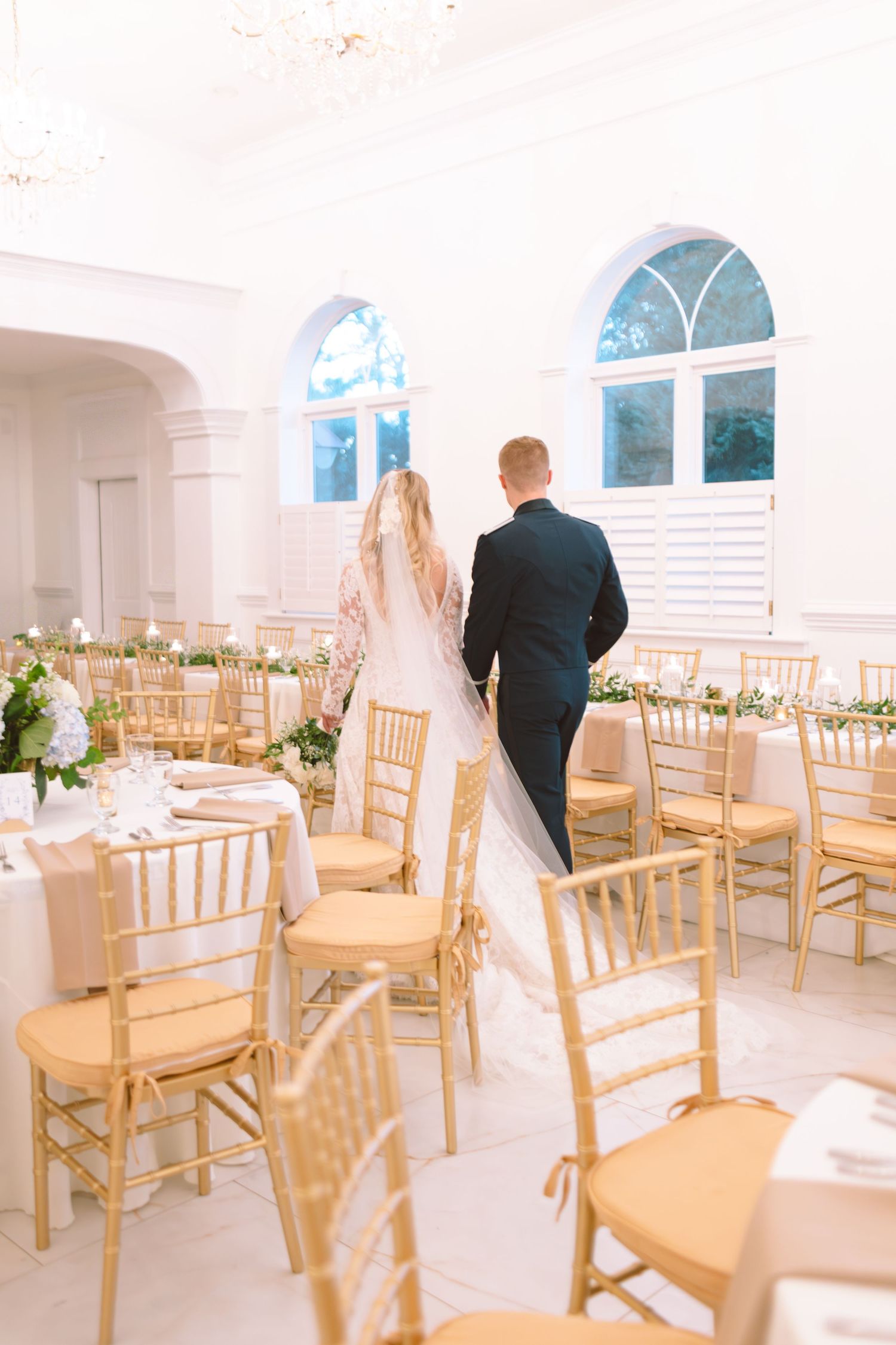 Bride and groom walking through a bright white elegant wedding reception hall with gold chiavari chairs.