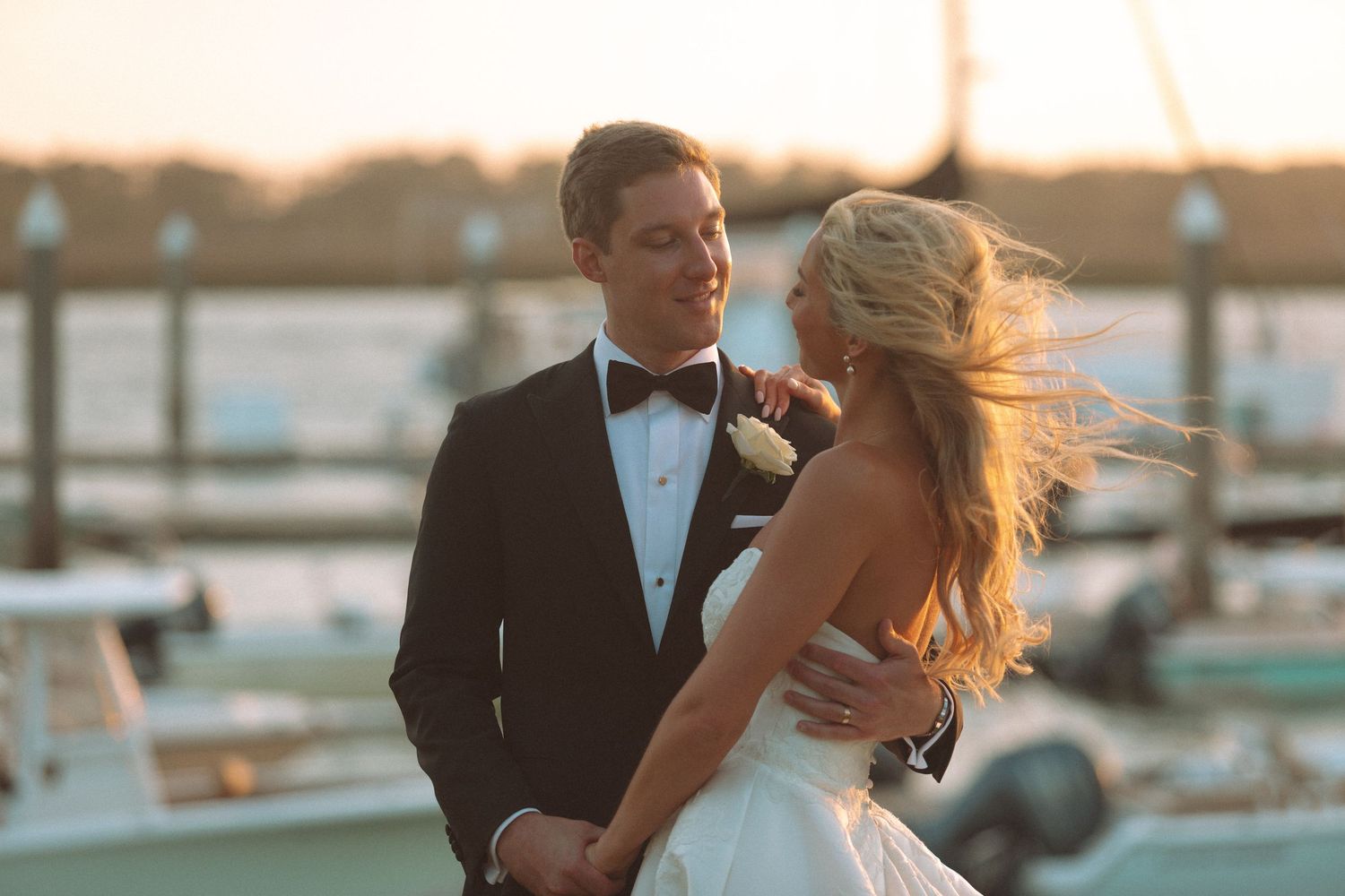 Bride and groom share romantic moment at waterfront marina during golden sunset.
