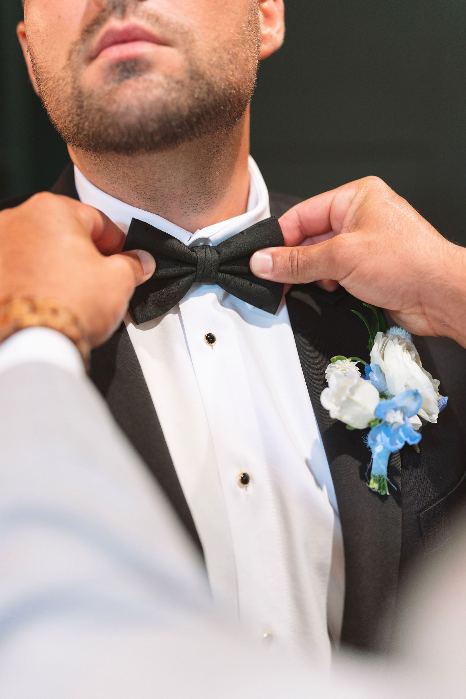 Close-up of hands adjusting a groom's black bow tie with a blue and white boutonniere on his lapel.