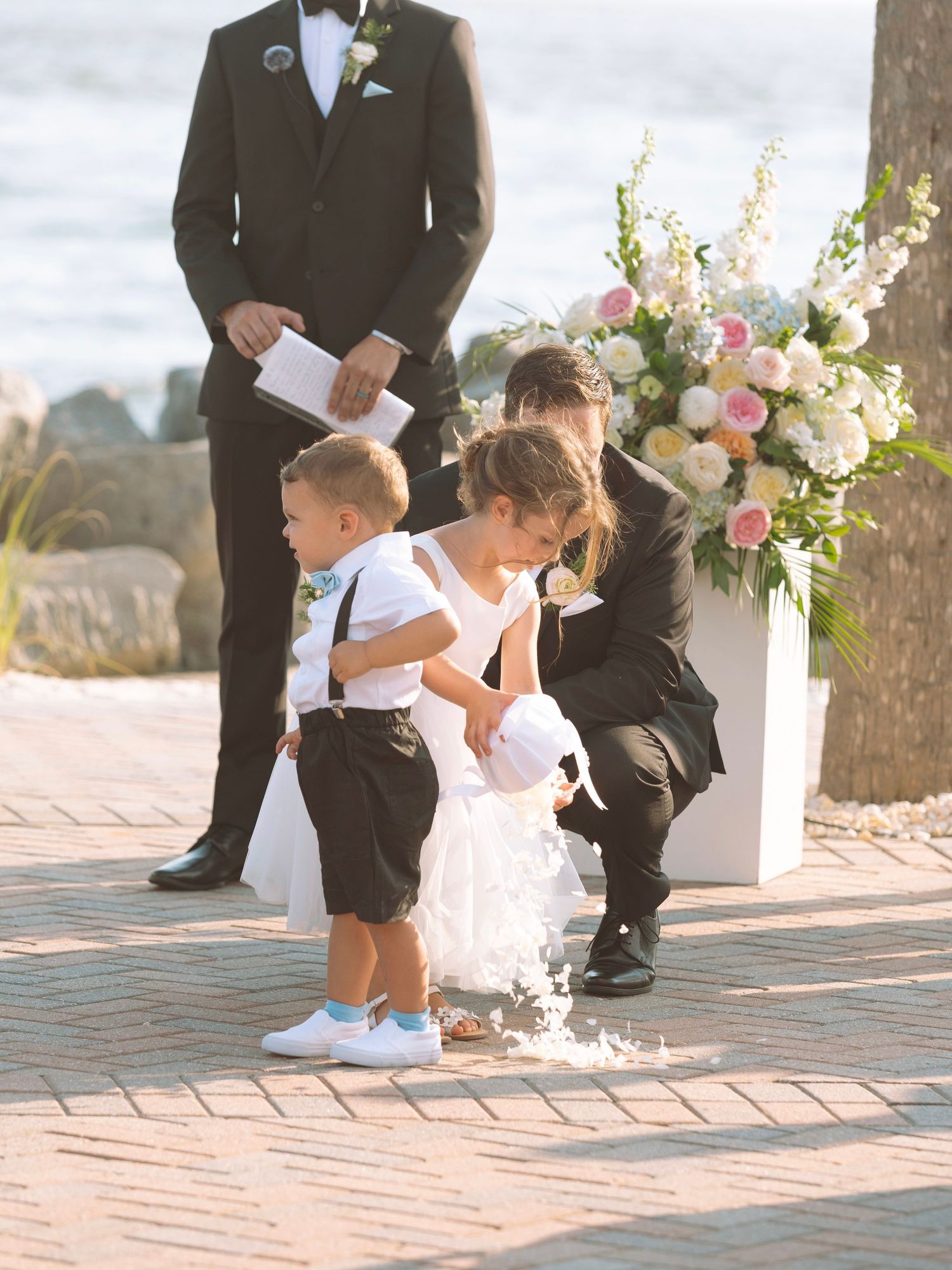 Flower girl and ring bearer at outdoor beach wedding ceremony with floral arrangements and officiant in background.