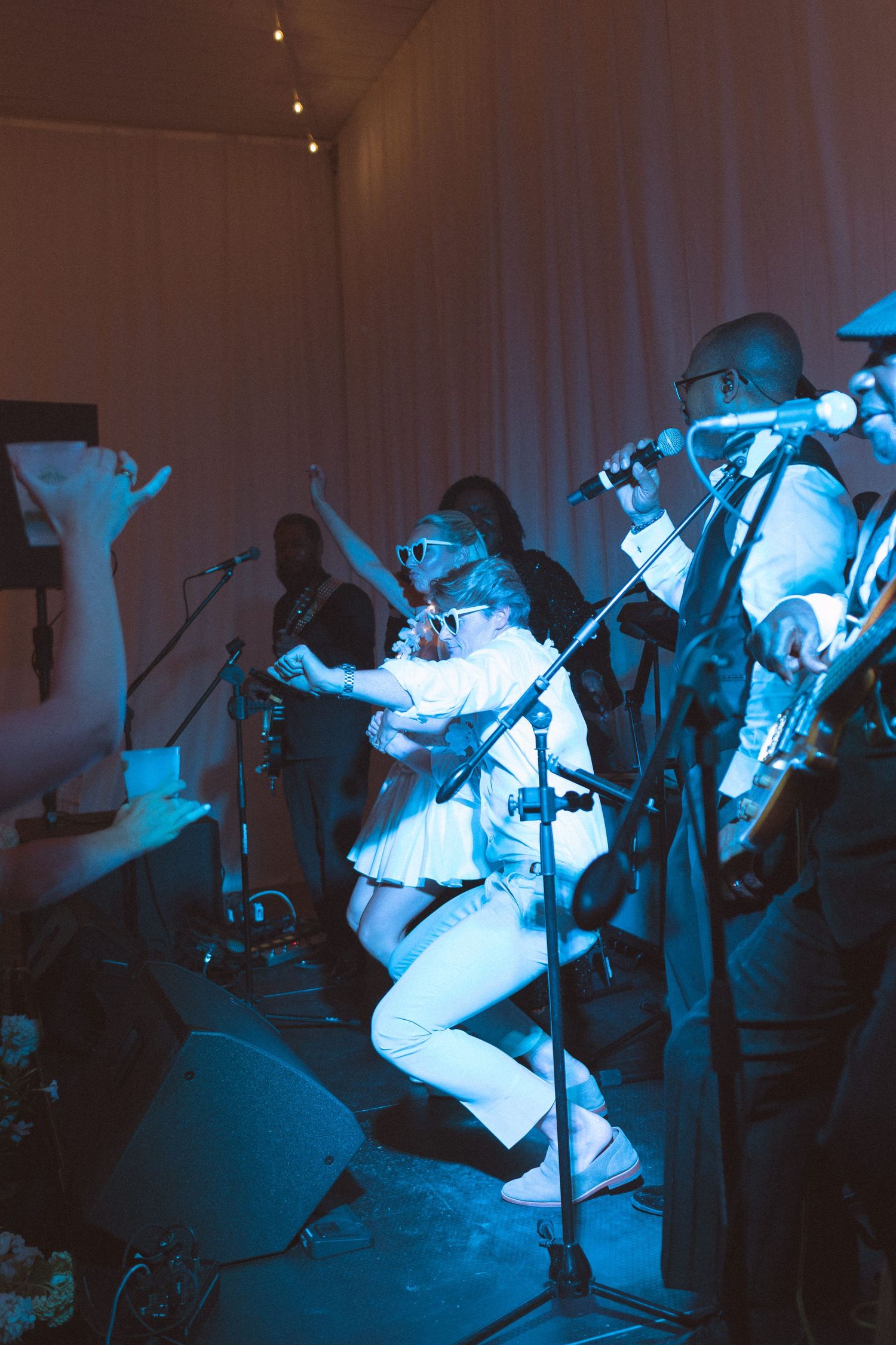 Live band performance under blue stage lights, musicians in white outfits playing instruments at an indoor event.