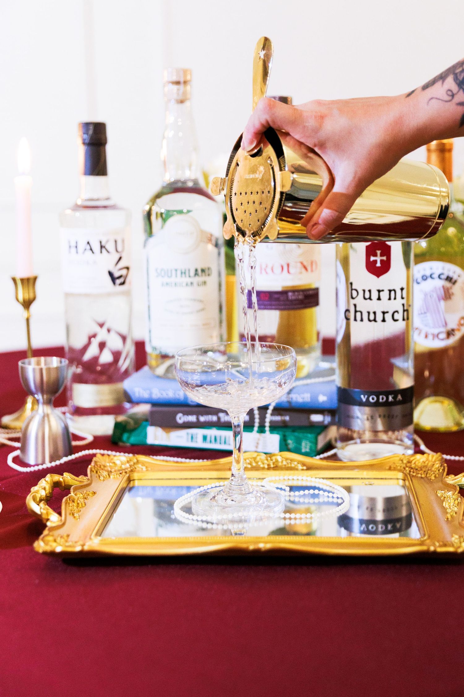 Bartender pouring cocktail through gold strainer into coupe glass on ornate tray with liquor bottles.