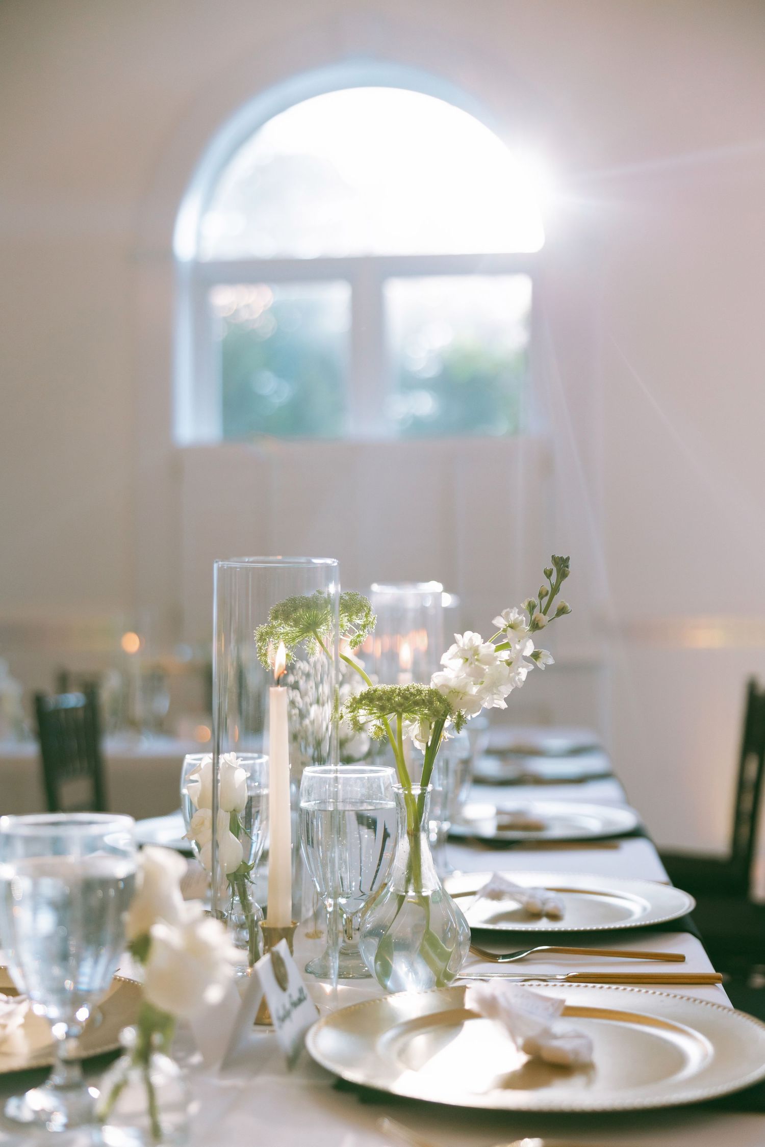 Elegant wedding reception table setting with white flowers, candles, and glassware in a bright sunlit venue.