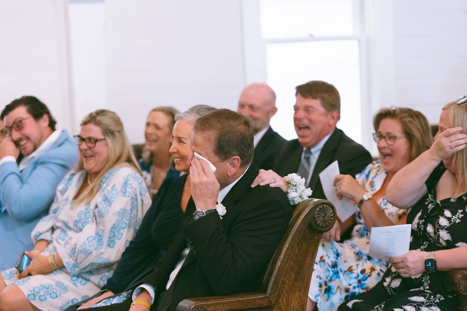 Wedding guests laughing and crying with joy during an emotional ceremony in a white chapel.