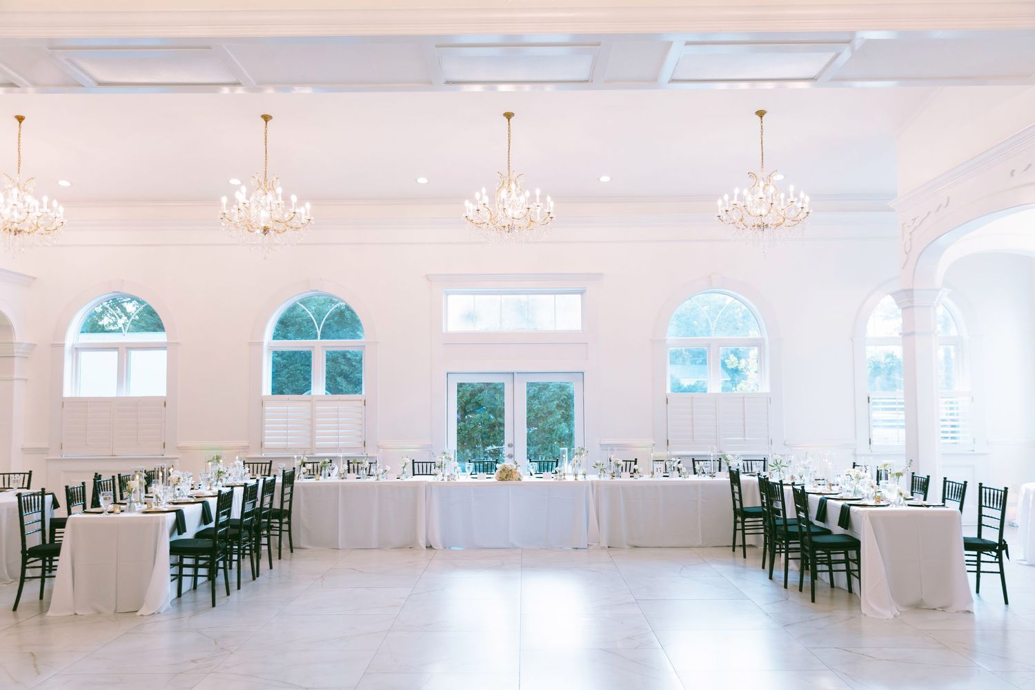 Elegant white ballroom wedding reception with chandeliers, arched windows, long tables, and dark chiavari chairs.
