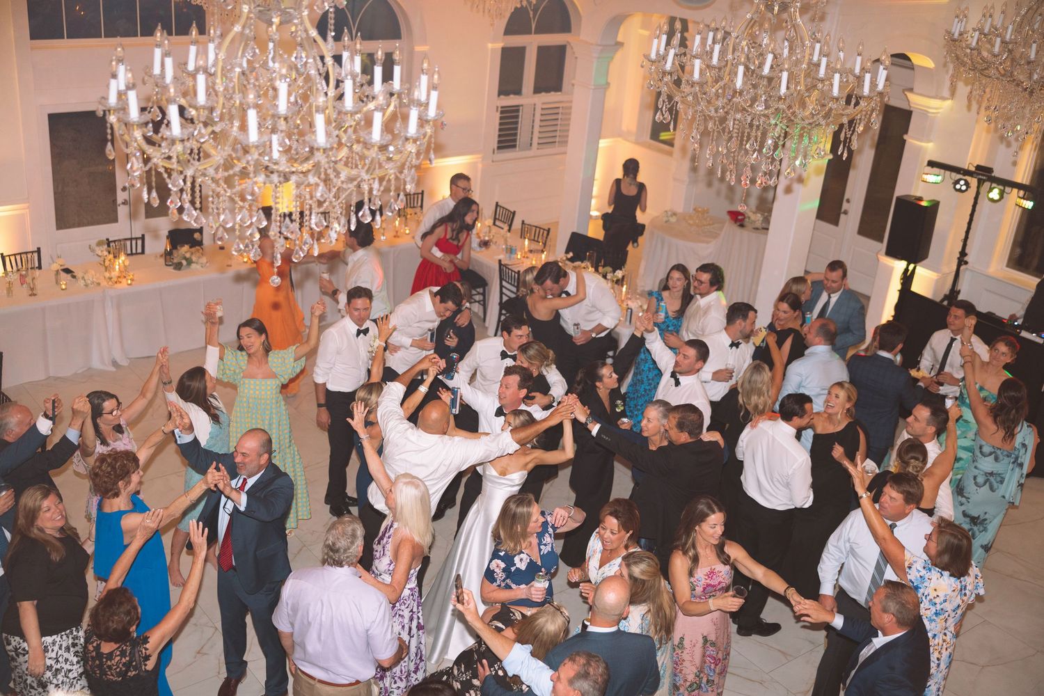 Elegant wedding reception with guests dancing under crystal chandeliers in a grand white ballroom.