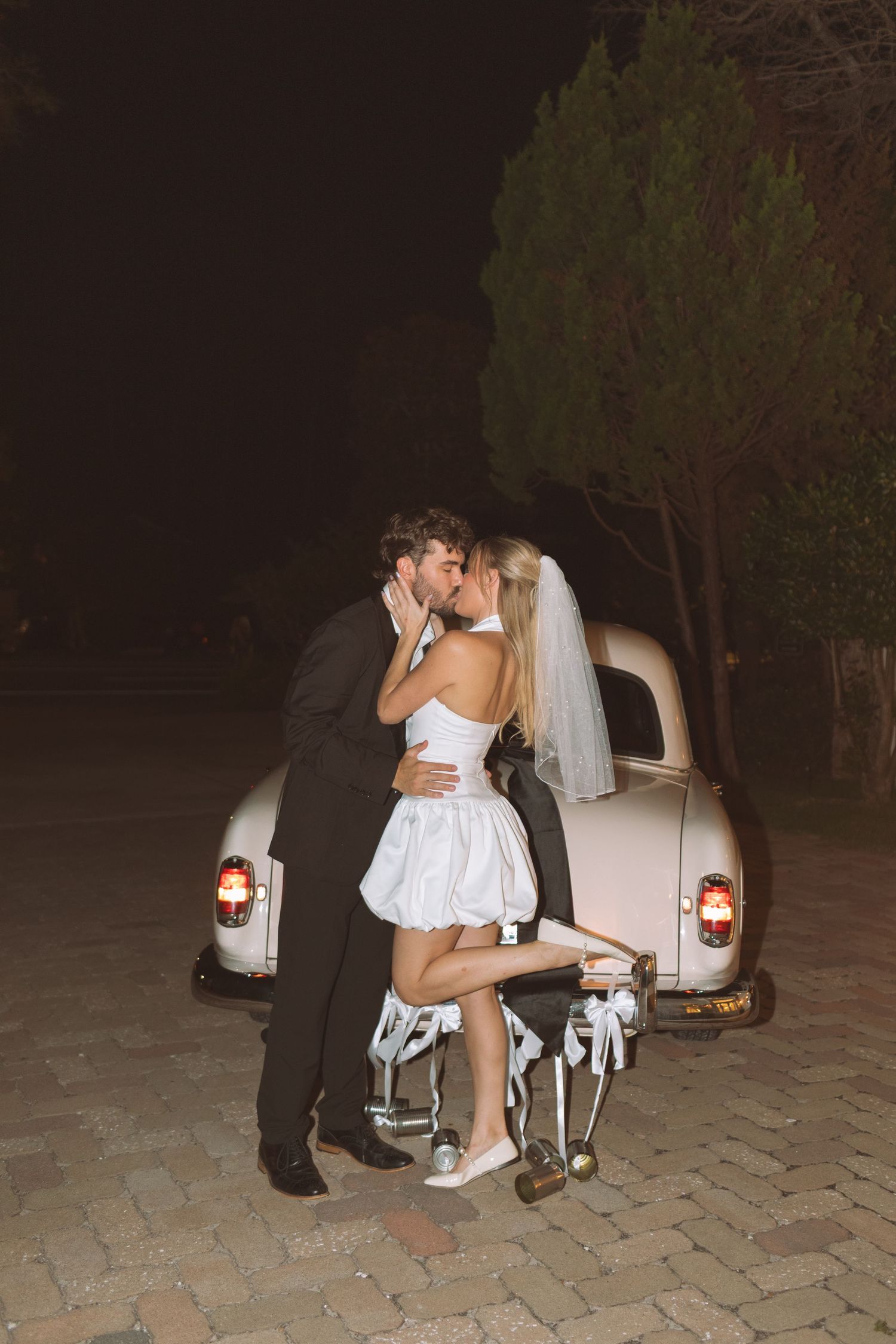 Newlywed couple sharing a romantic kiss beside a vintage white car at night, bride in short white dress and veil.