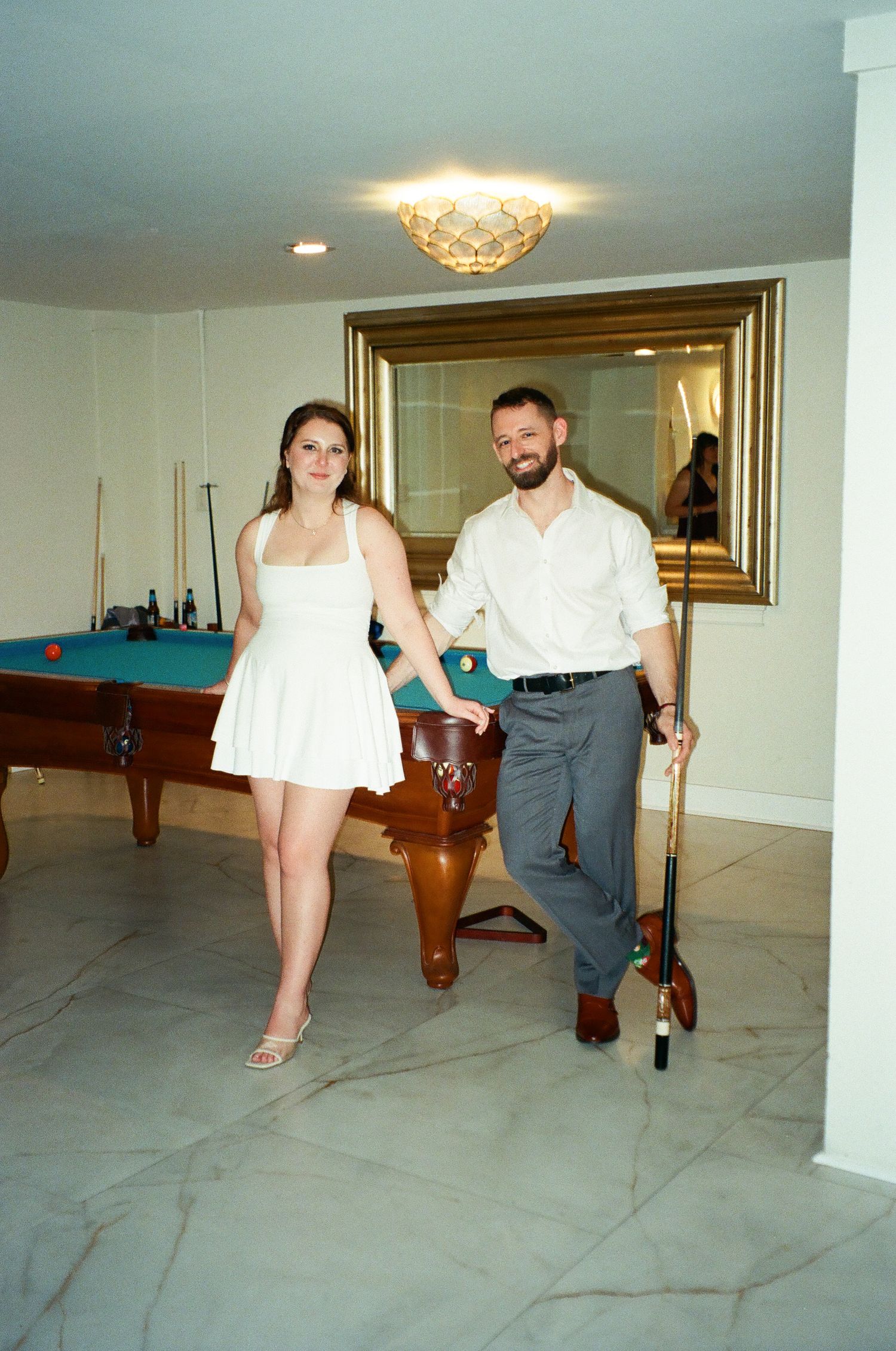 A smiling couple holds hands near a pool table in a game room, woman in white dress, man in white shirt holding a cue.