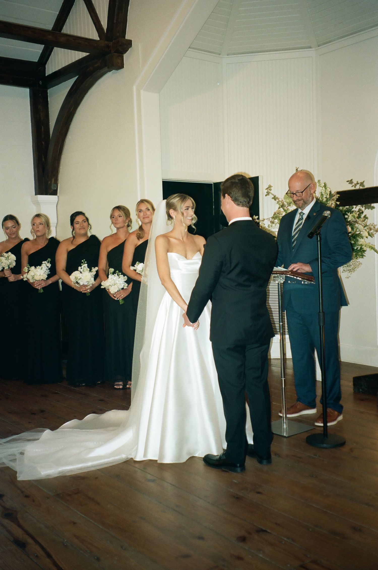 Bride and groom hold hands at indoor wedding ceremony with bridesmaids in black gowns holding white bouquets.