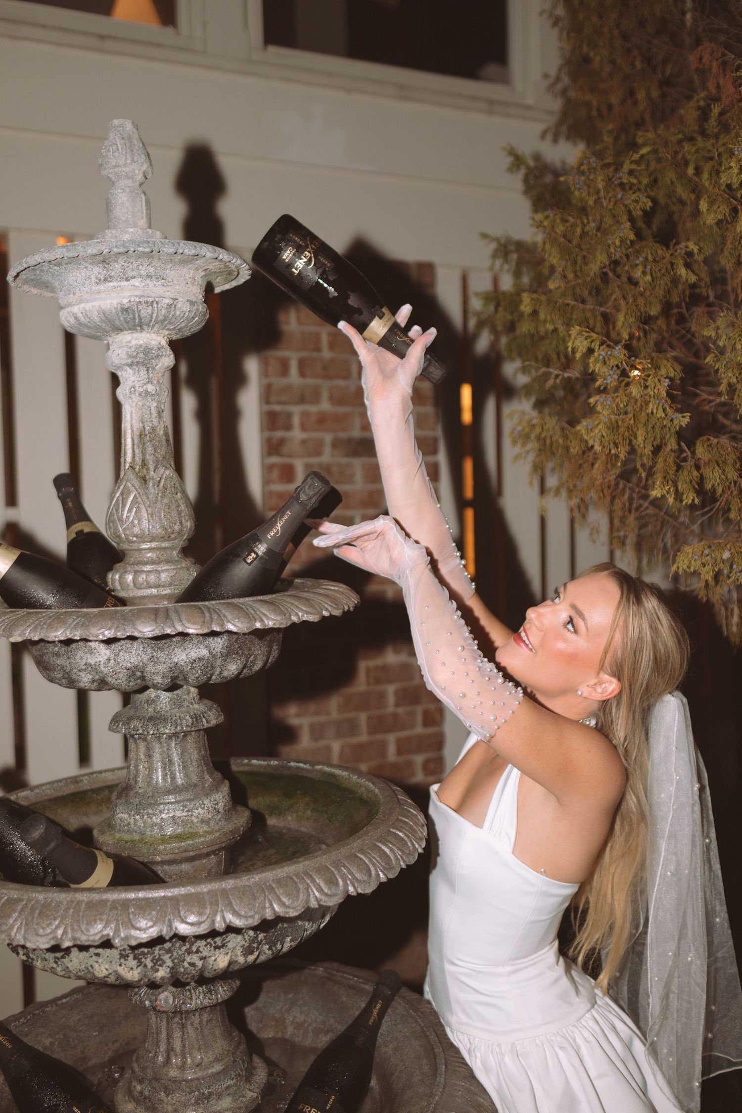 Bride in white wedding dress pouring champagne into an outdoor stone fountain at night.