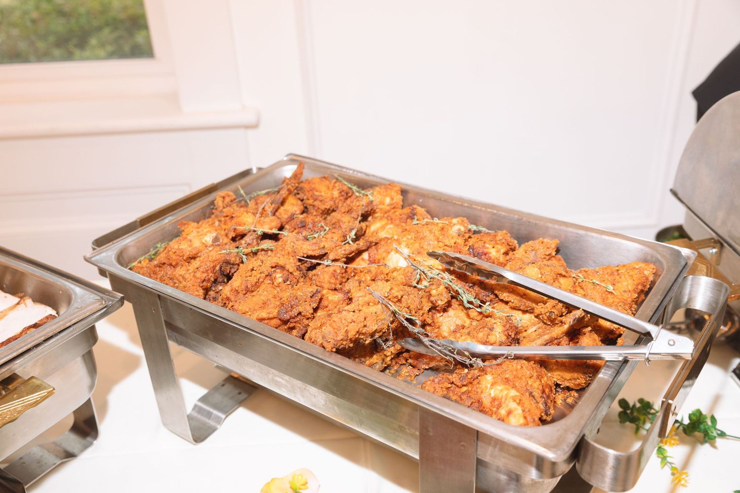 Crispy fried chicken pieces in a silver chafing dish with tongs at a buffet.