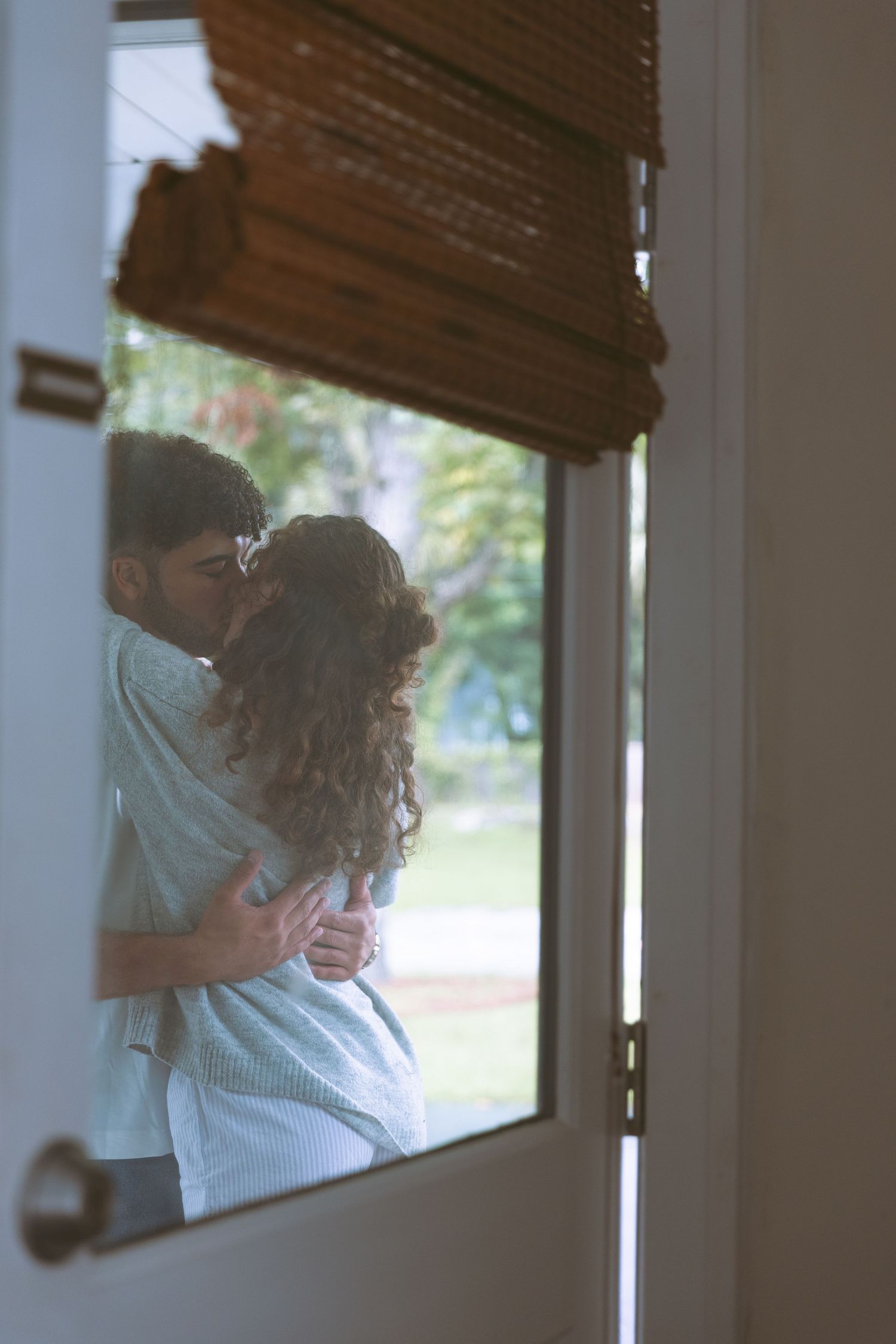 Couple sharing tender kiss and embrace seen through window with wooden blinds.
