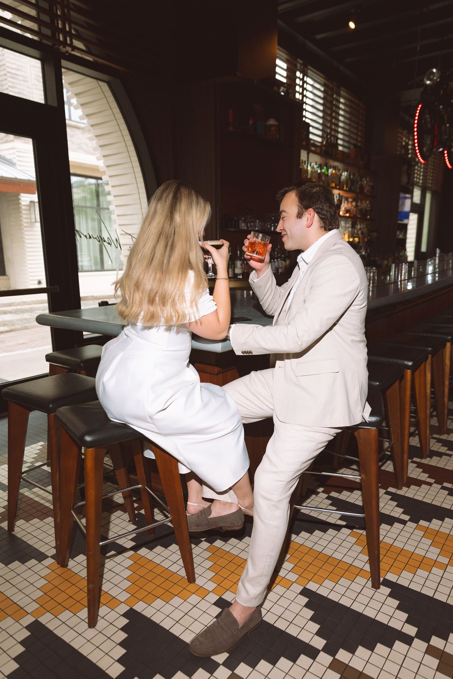 Elegant couple in white outfits enjoying cocktails at a stylish bar, sitting on bar stools in a sophisticated restaurant setting.