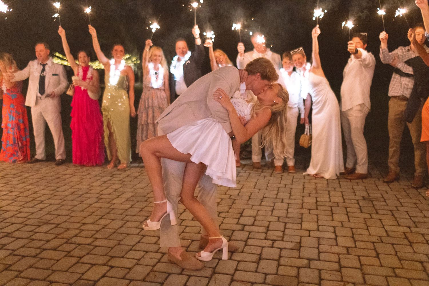 Groom dipping bride during sparkler send-off surrounded by cheering wedding guests.