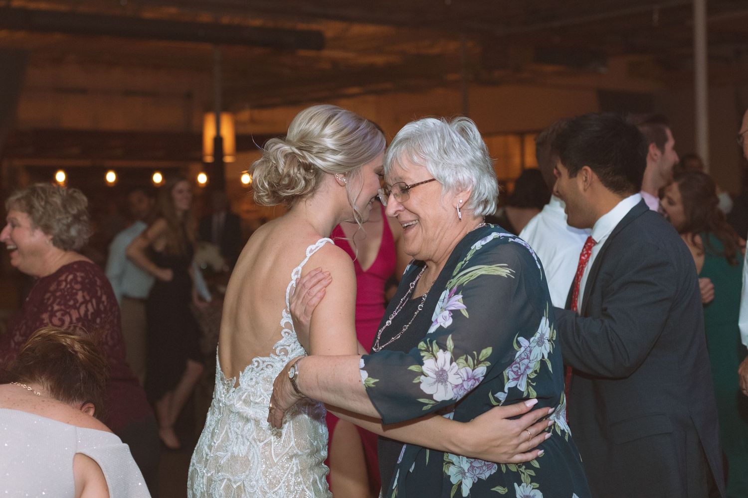 Bride sharing a warm dance with an elderly woman guest at a rustic wedding reception.