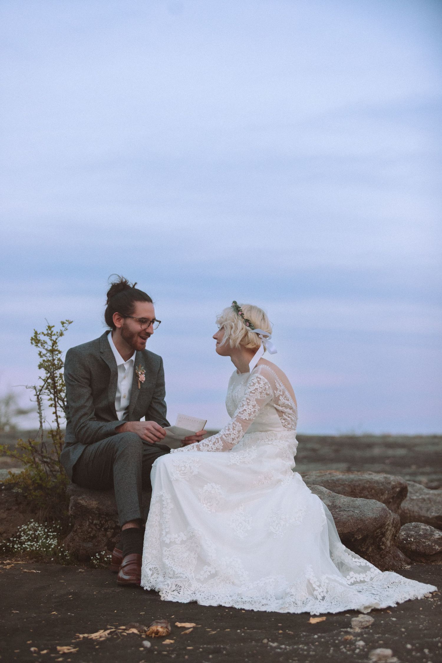 A bride and groom sit together on rocks at dusk, smiling at each other in wedding attire.