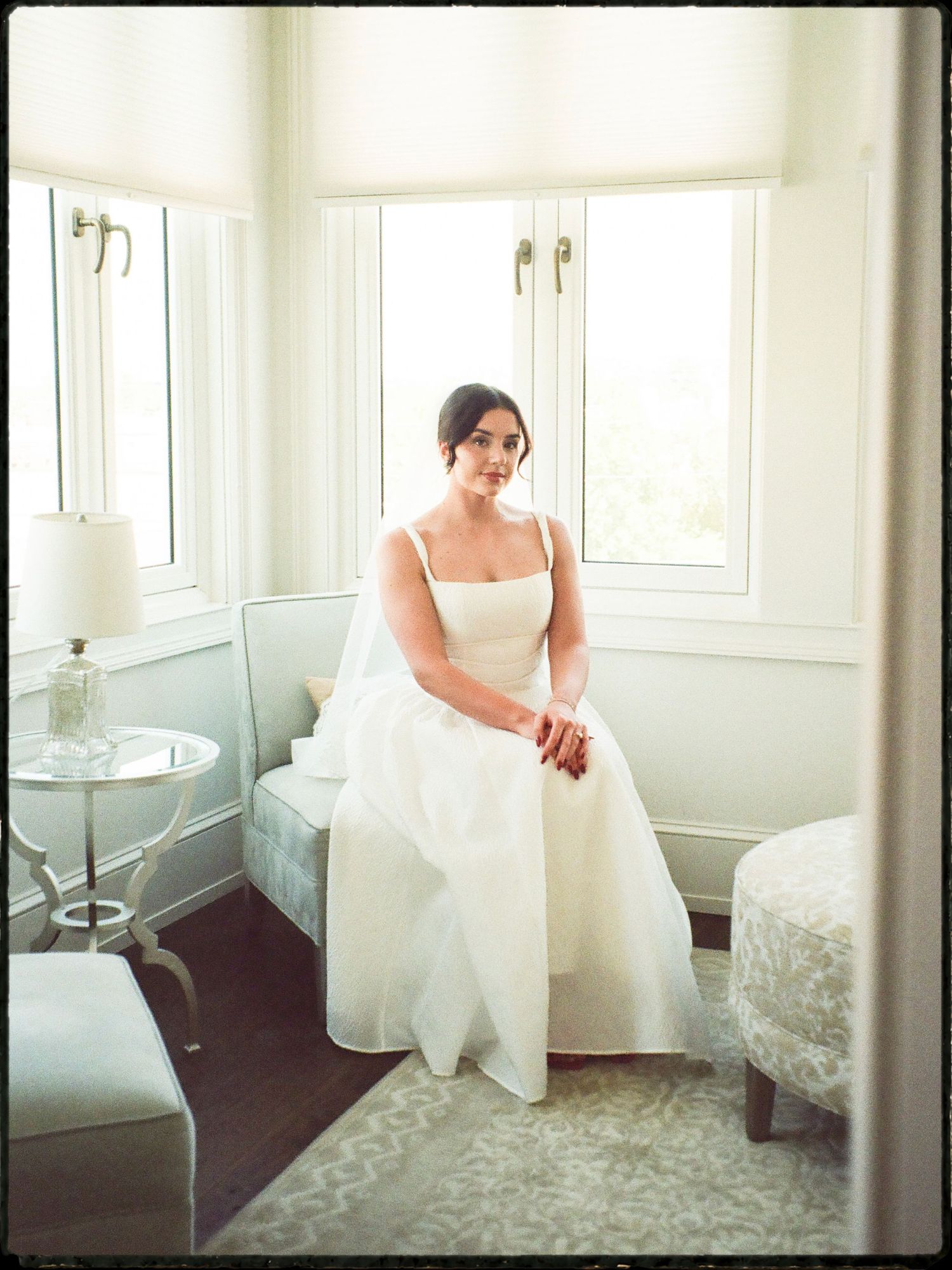 Elegant bride in white gown sitting by bright windows in a softly lit bridal suite.