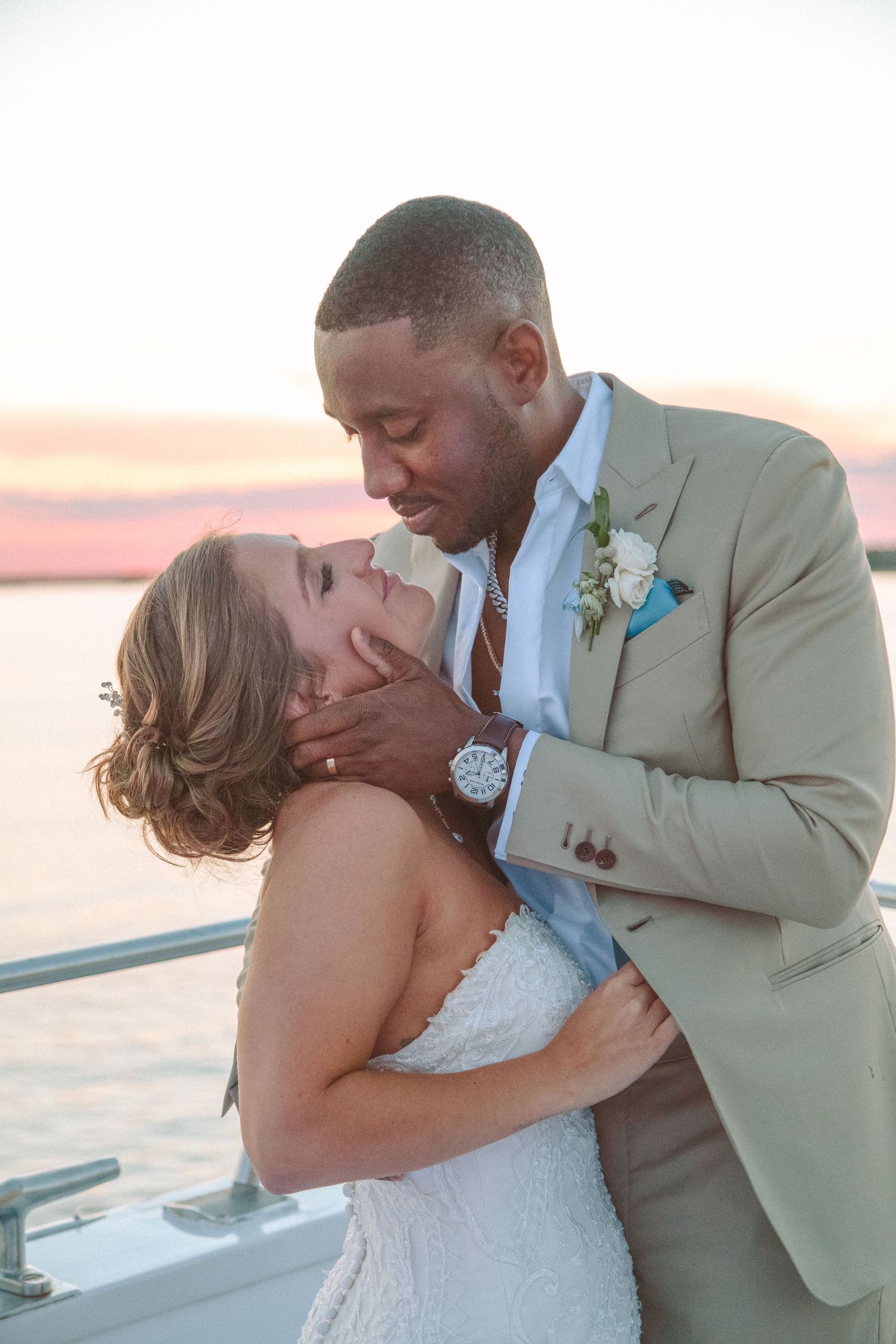 Groom in tan suit tenderly holds bride's face during romantic sunset portrait on a boat deck.