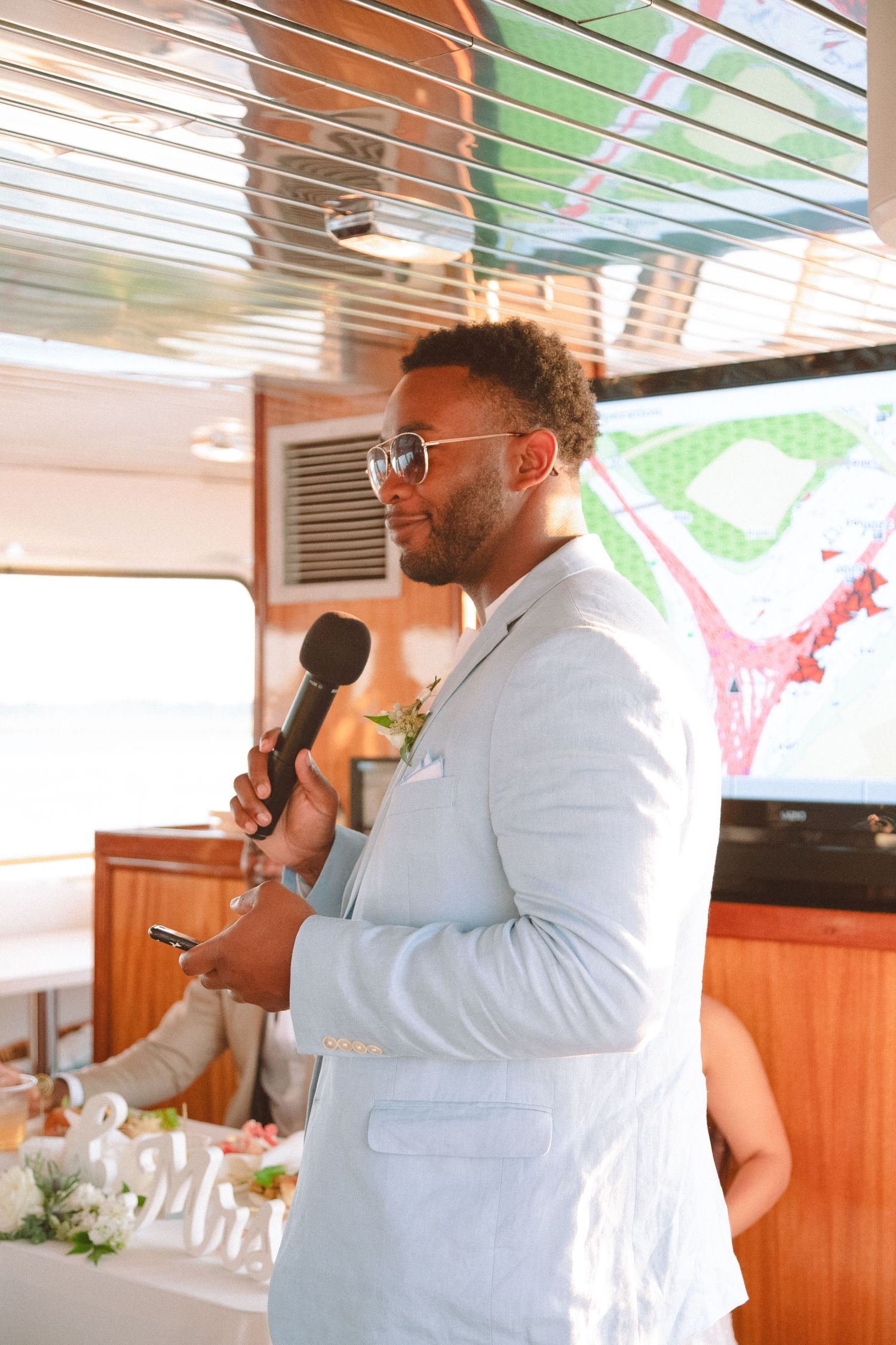 Groomsman in light gray suit giving a wedding speech while holding a microphone on a boat reception.