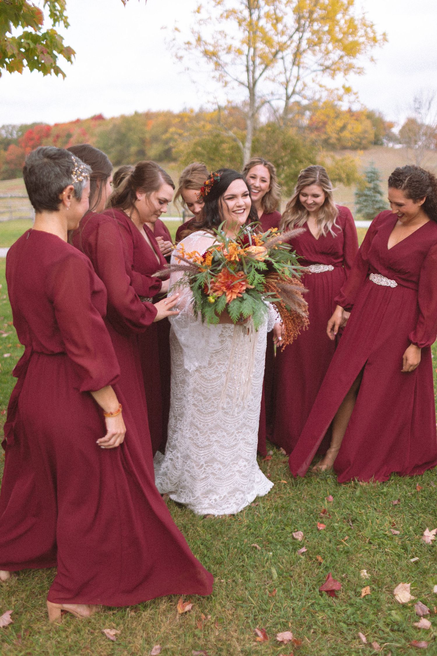 Bride in lace gown surrounded by bridesmaids in burgundy dresses holding autumn bouquet outdoors.